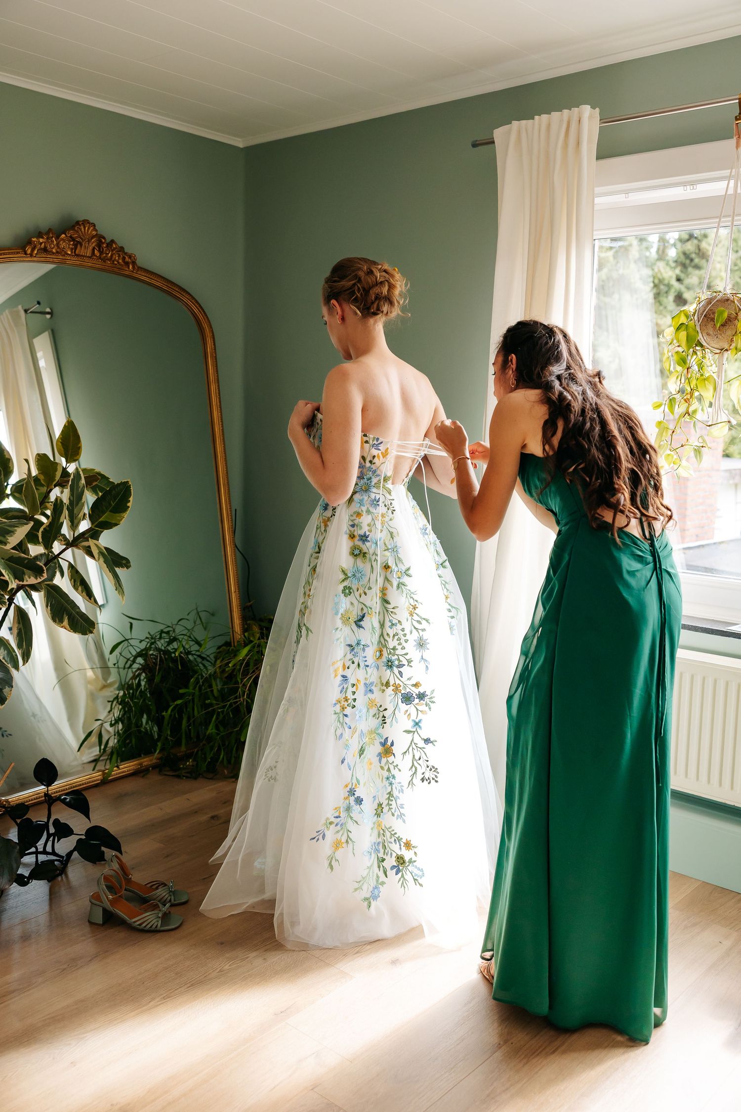 Bridesmaid in emerald green dress lacing up bride's floral embroidered wedding gown by window.