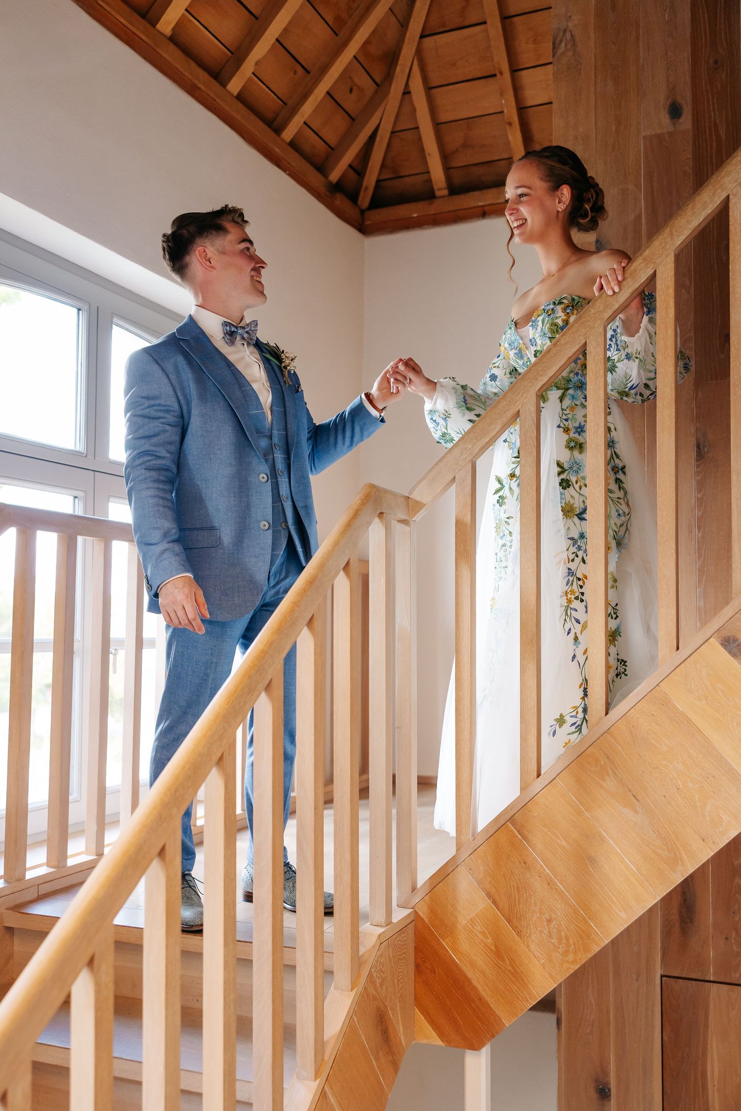 Smiling bride and groom in blue suit holding hands on wooden staircase inside wedding venue.