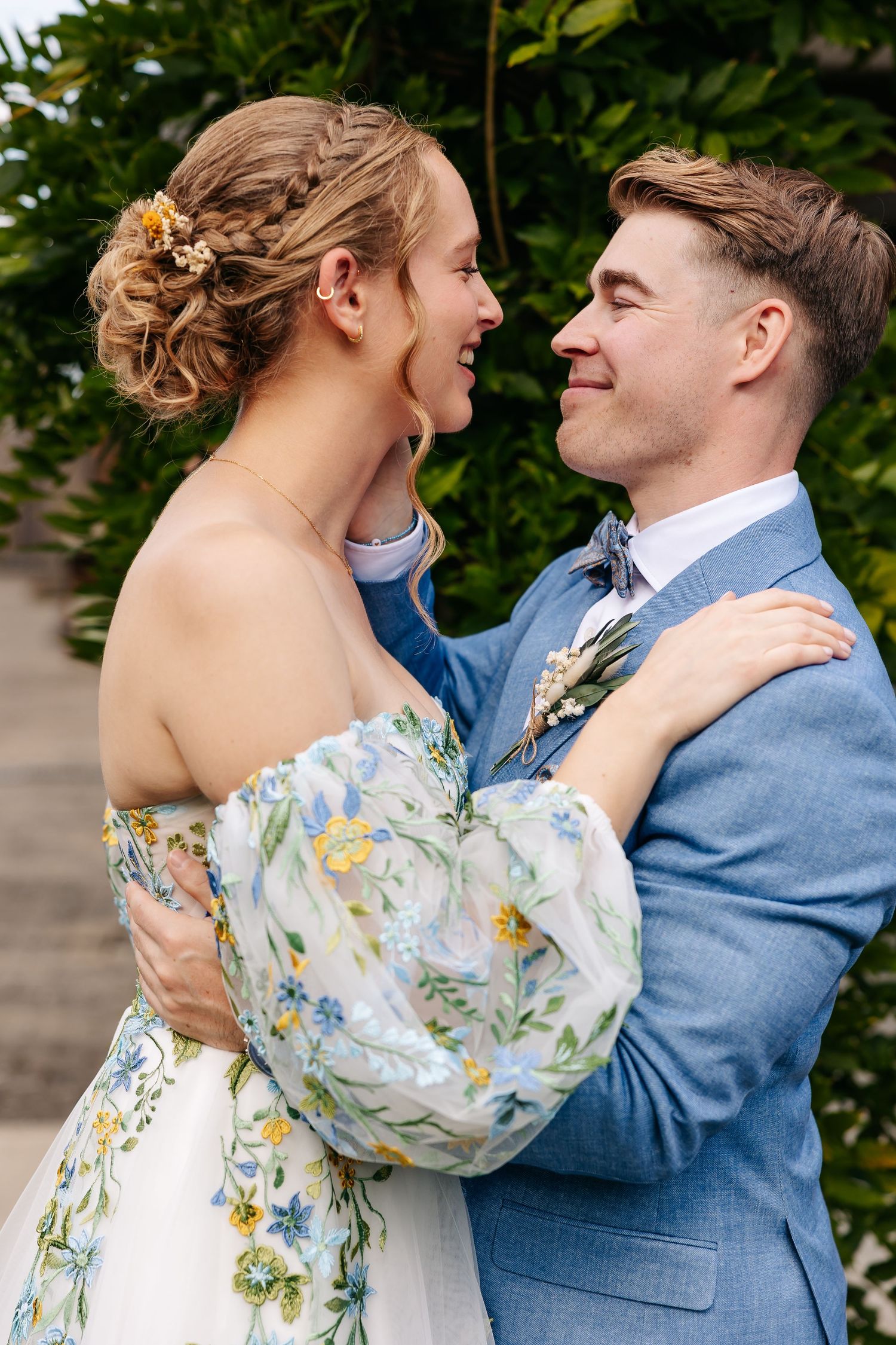 Bride in floral dress and groom in blue suit embrace and laugh together outdoors on their wedding day.