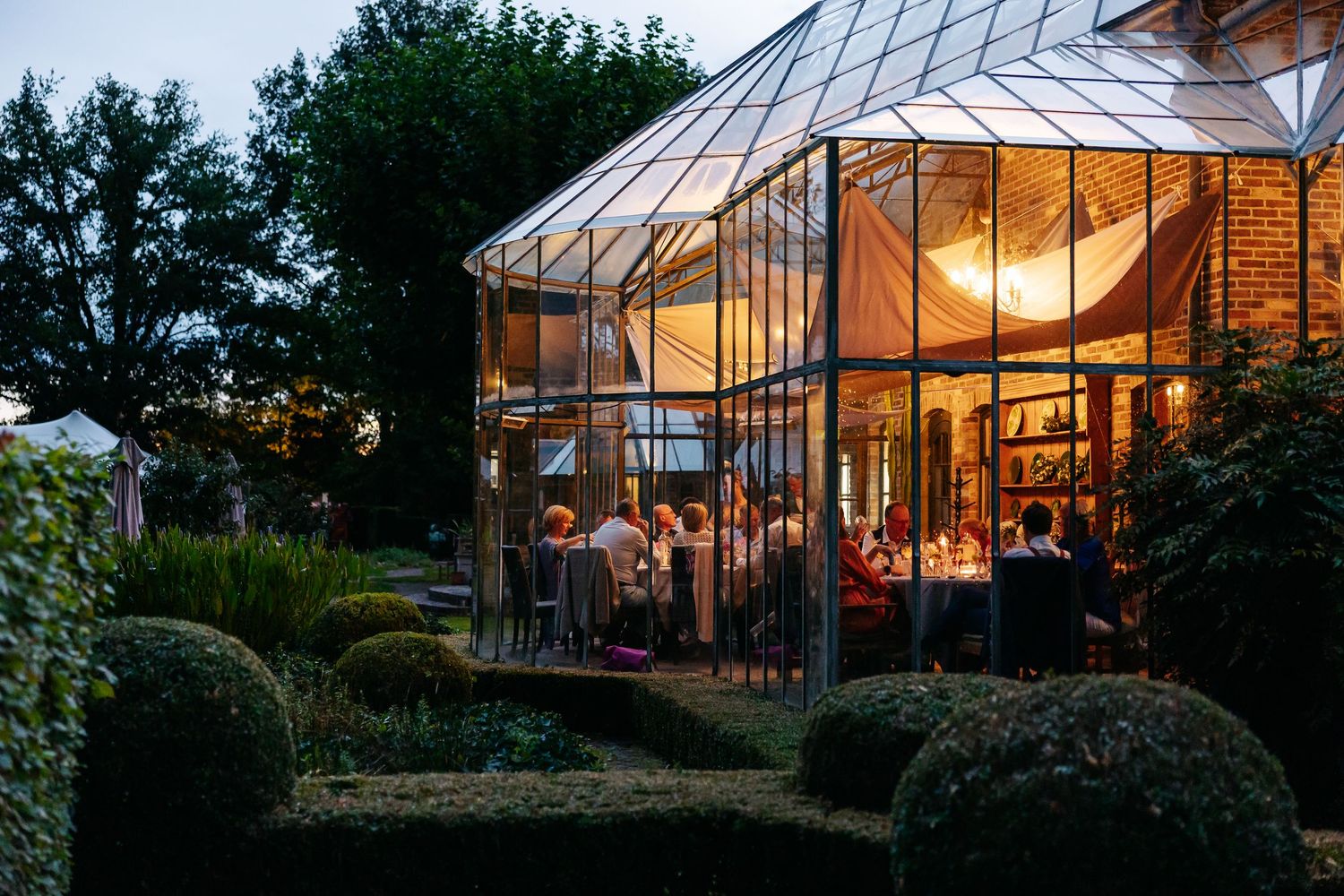 Elegant glass greenhouse wedding reception glowing warmly at twilight with guests dining inside.