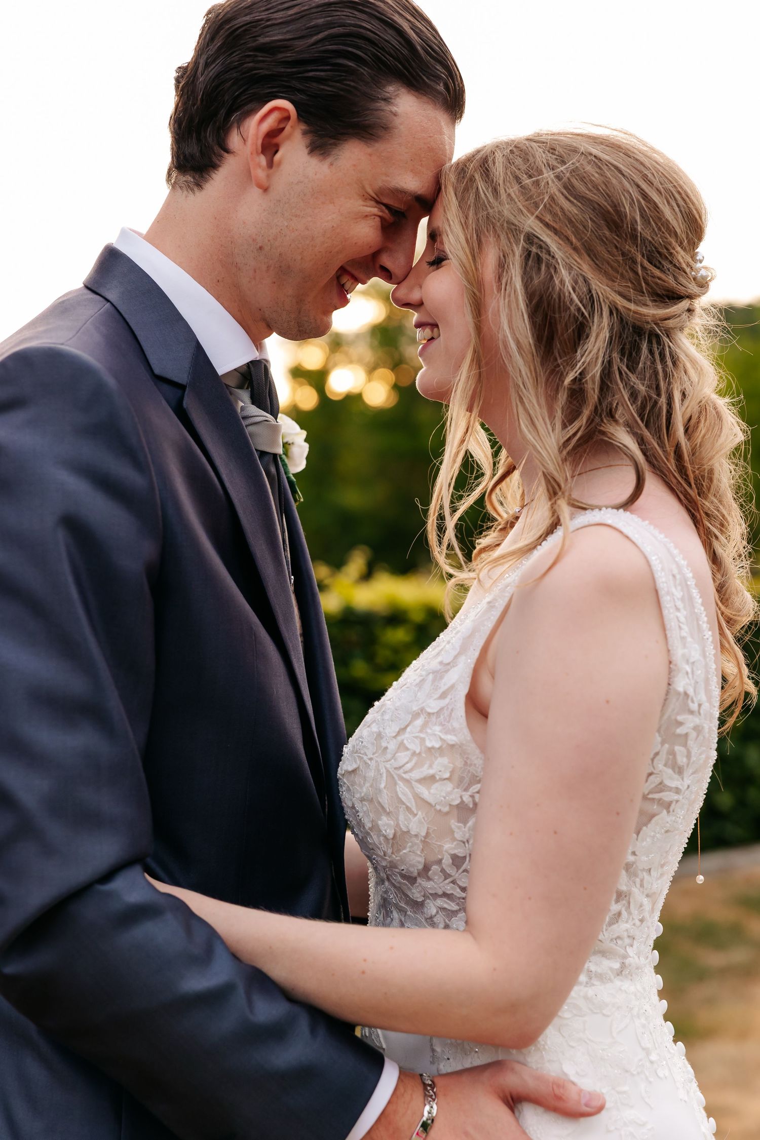 Bride and groom touch foreheads and smile warmly in a golden sunset garden wedding portrait.