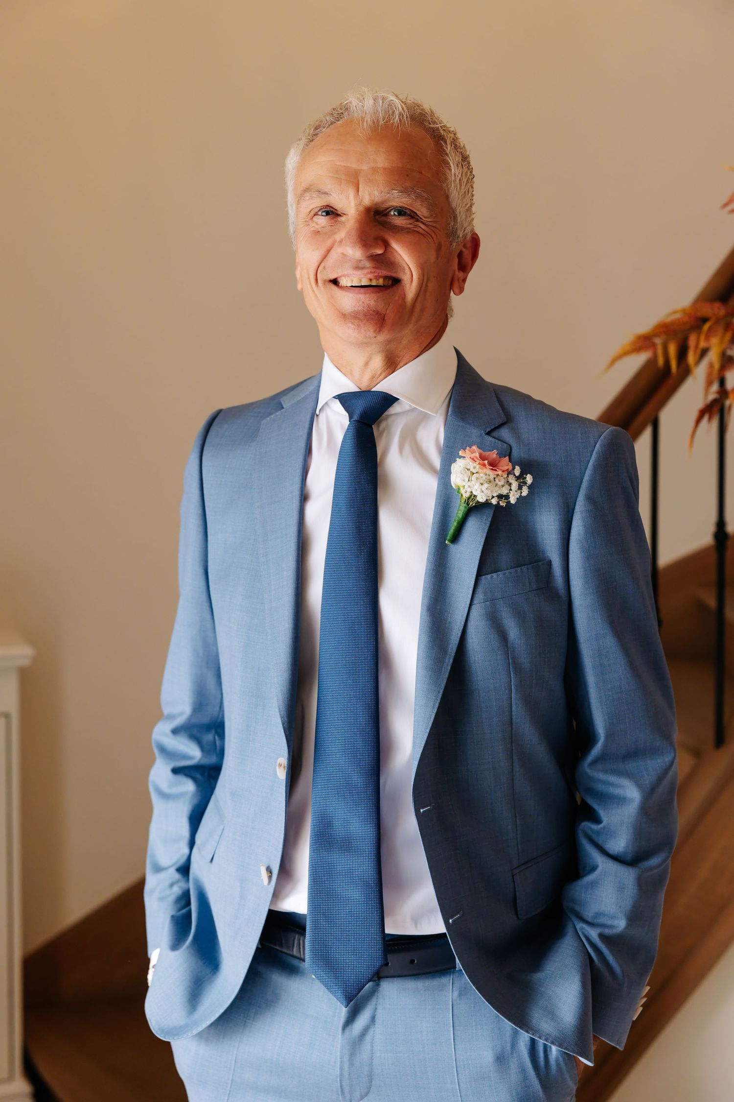 Dapper groom in blue suit with floral boutonniere smiles warmly while standing near a wooden staircase.