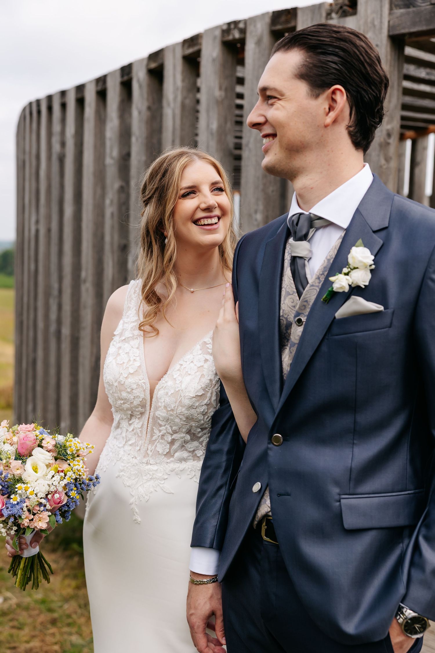 Laughing bride in lace gown and groom in navy suit share a joyful moment outdoors on wedding day.