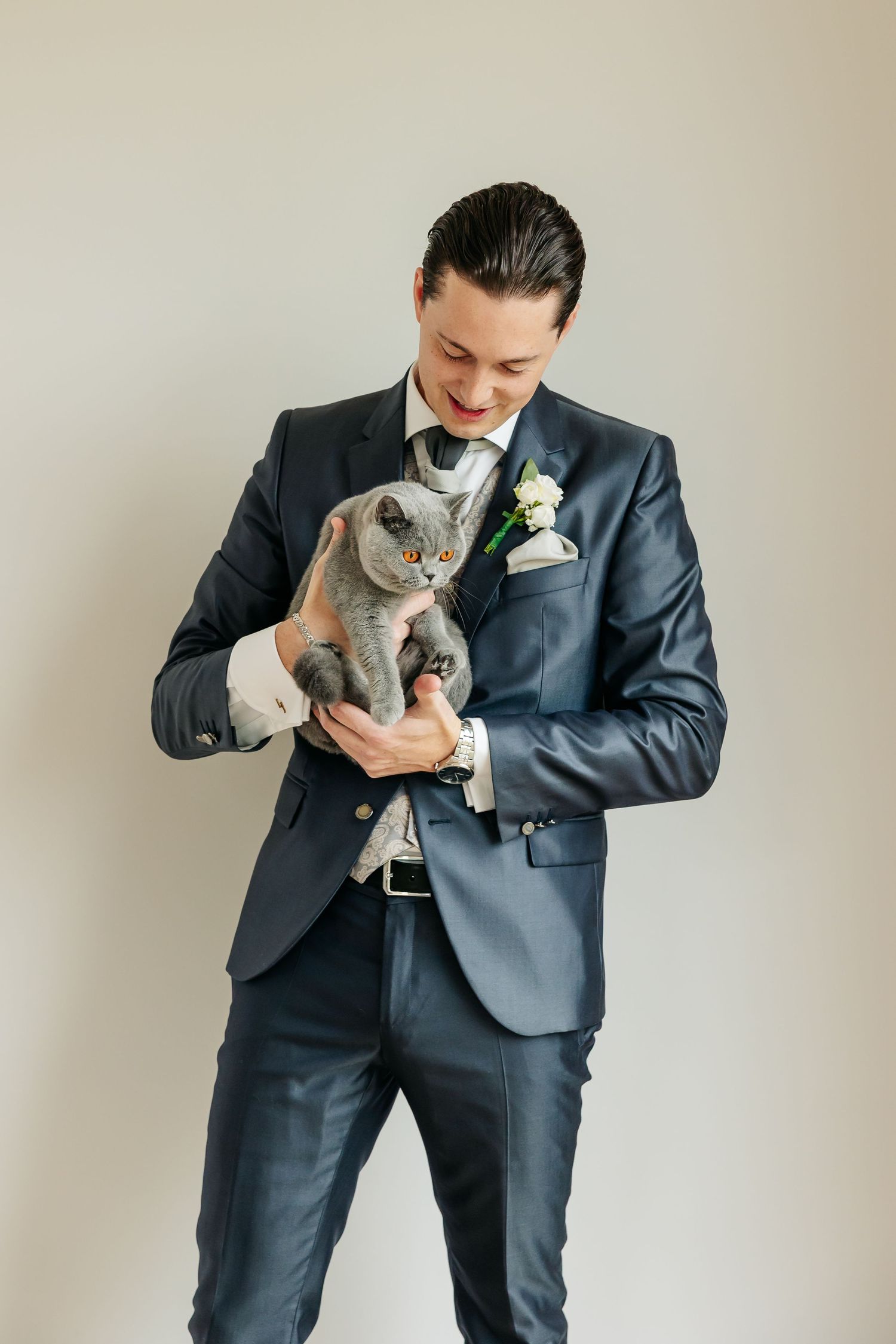Smiling groom in navy wedding suit holds a gray British Shorthair cat against neutral background.