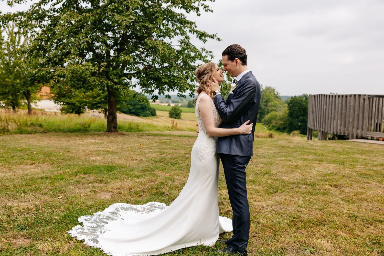 Bride and groom share a tender moment outdoors on green grass, with trees and a barn in the background.