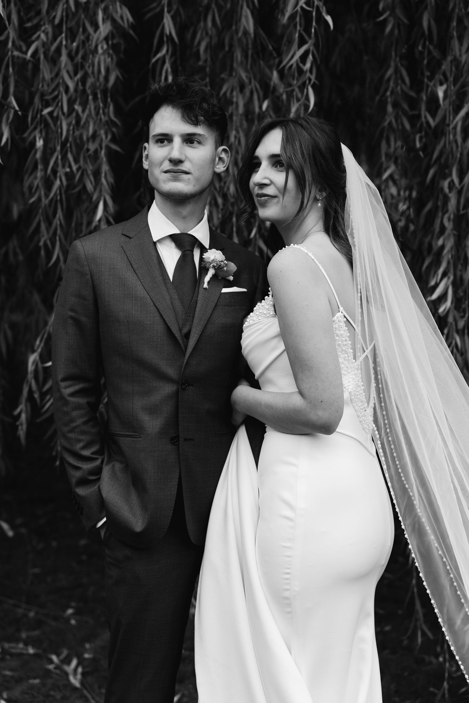 Black and white photo of bride in white gown and veil with groom in dark suit under willow tree.