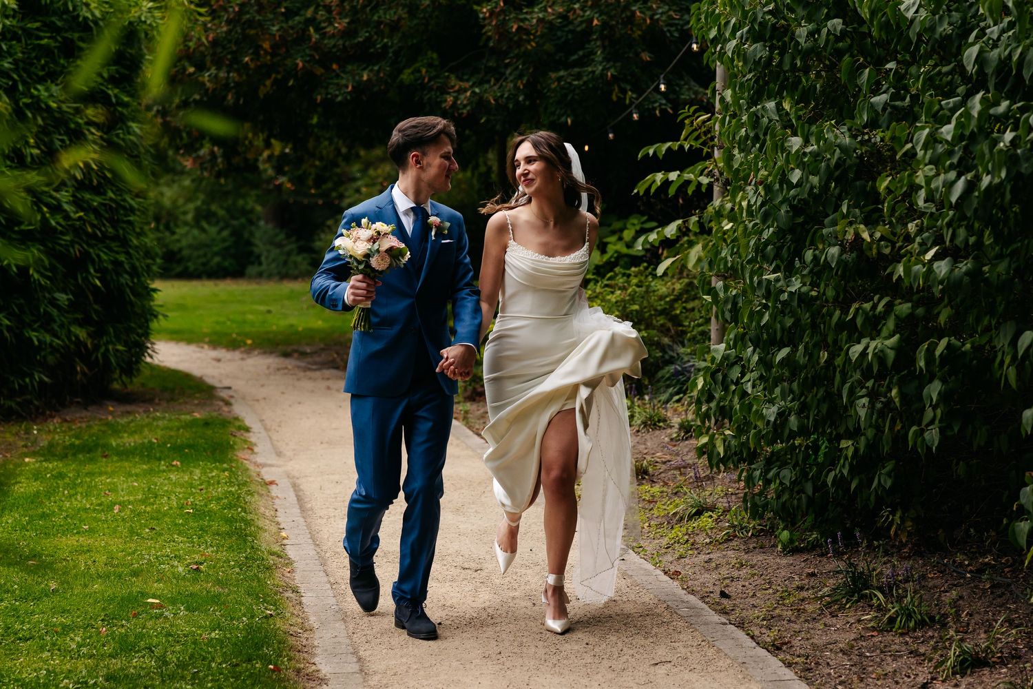 Bride and groom walking hand in hand along a lush garden path on their sunny wedding day.