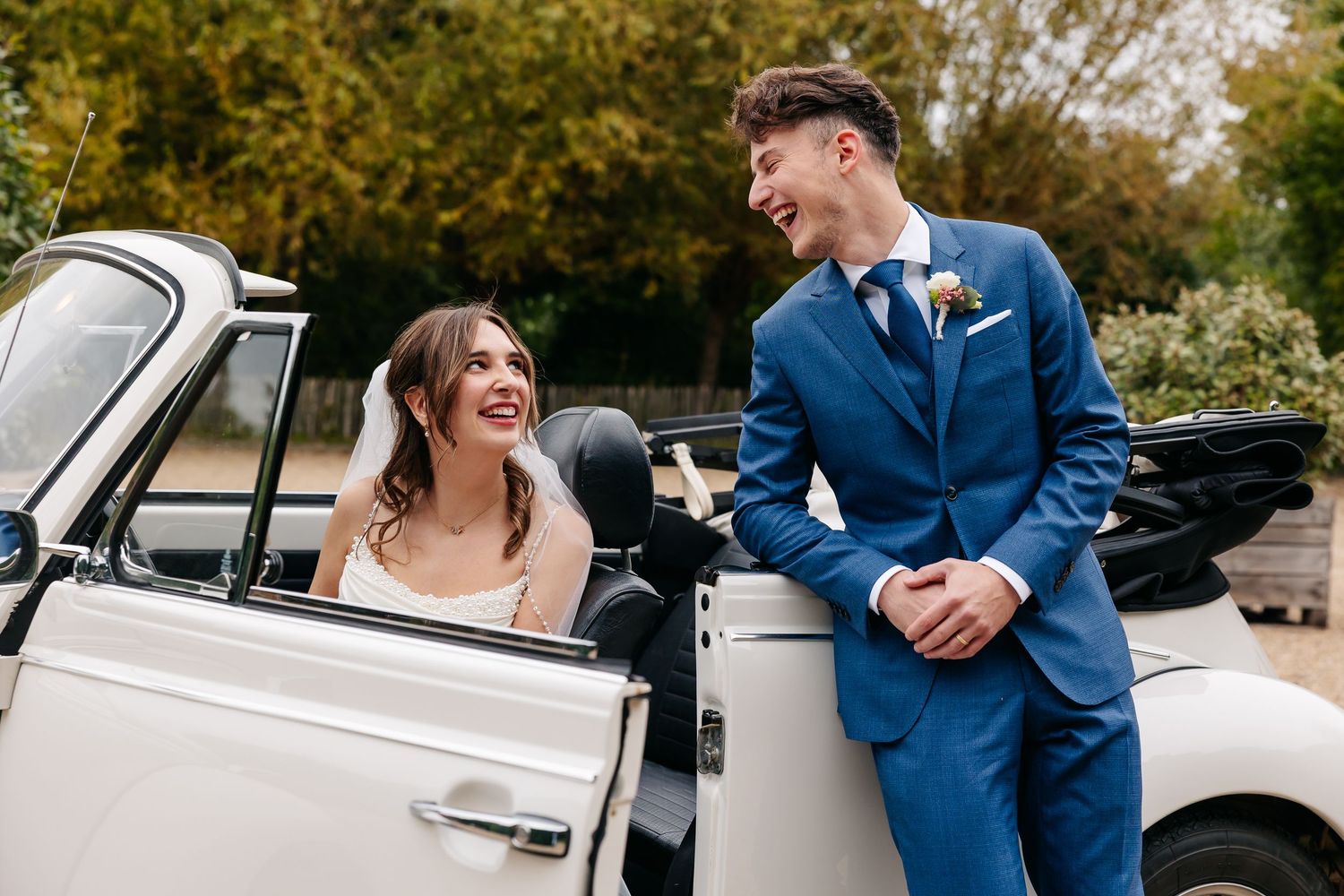 Smiling bride and groom in blue suit sharing a joyful moment leaning against a white vintage wedding car.