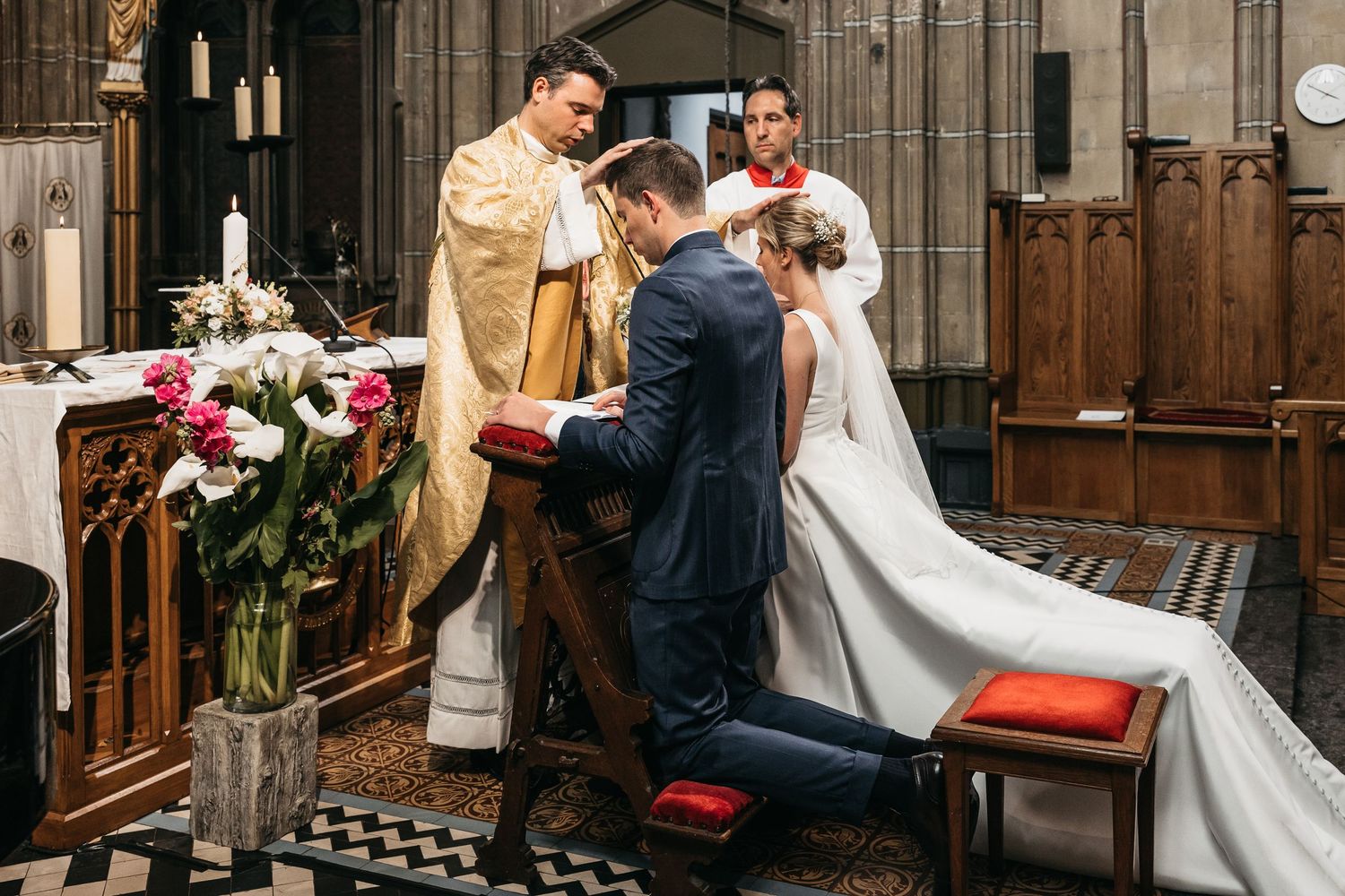 Couple kneeling at altar receiving blessing from priest during traditional Catholic church wedding.