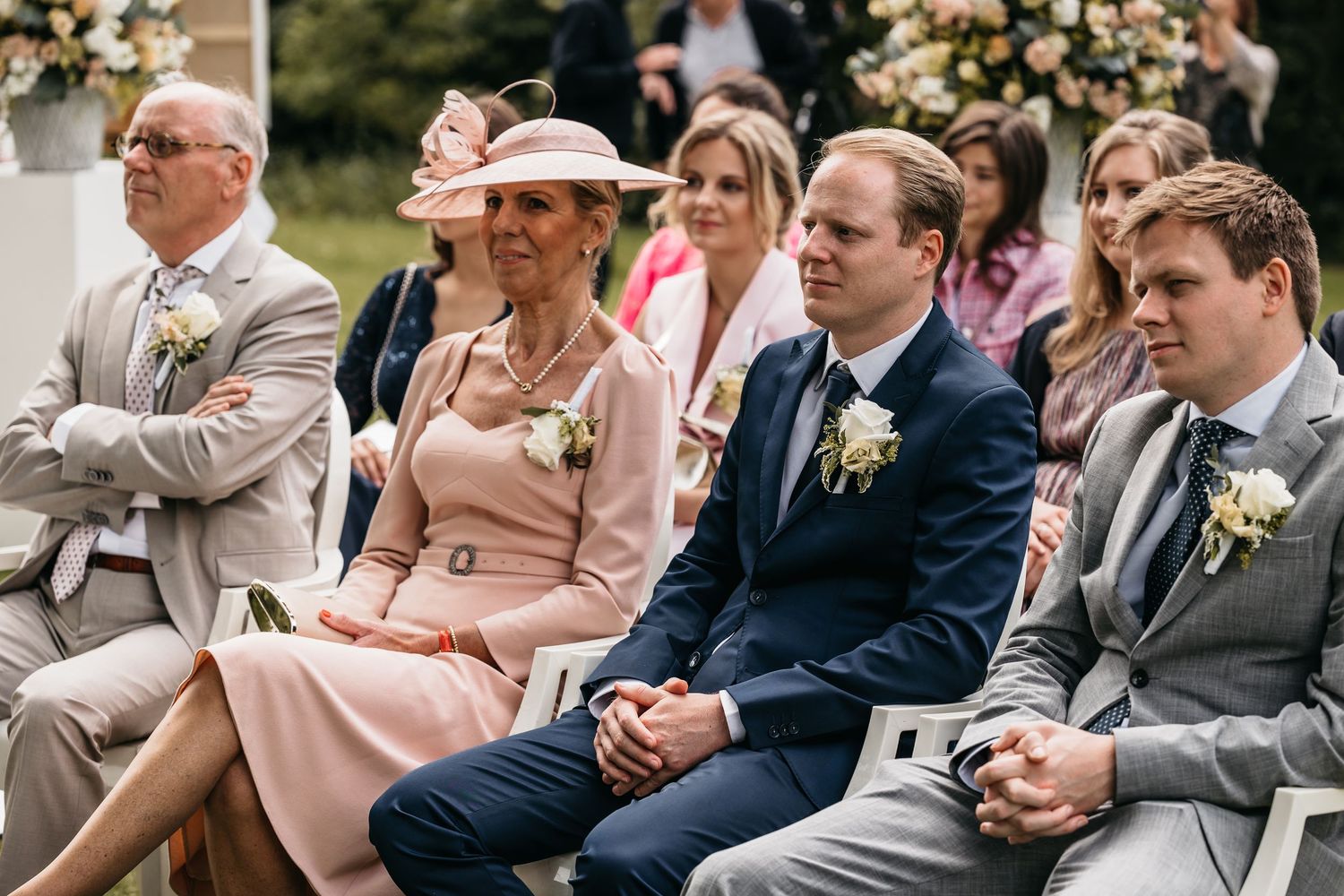 Elegantly dressed wedding guests seated at outdoor ceremony including woman in pink dress and fascinator hat.