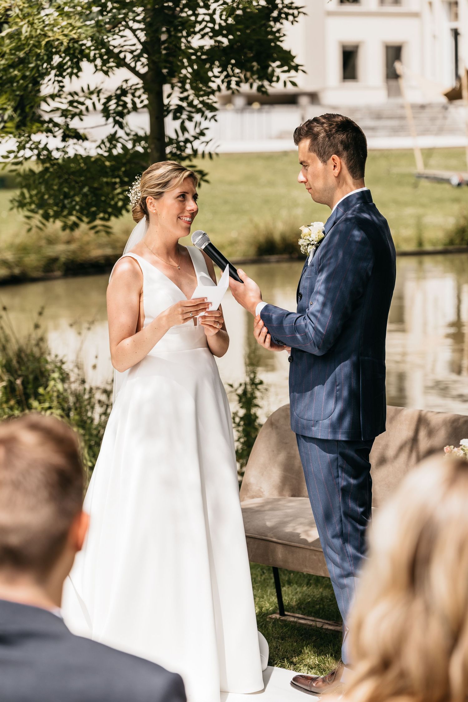 Bride reads wedding vows into microphone while groom holds her hand during outdoor ceremony by a pond.