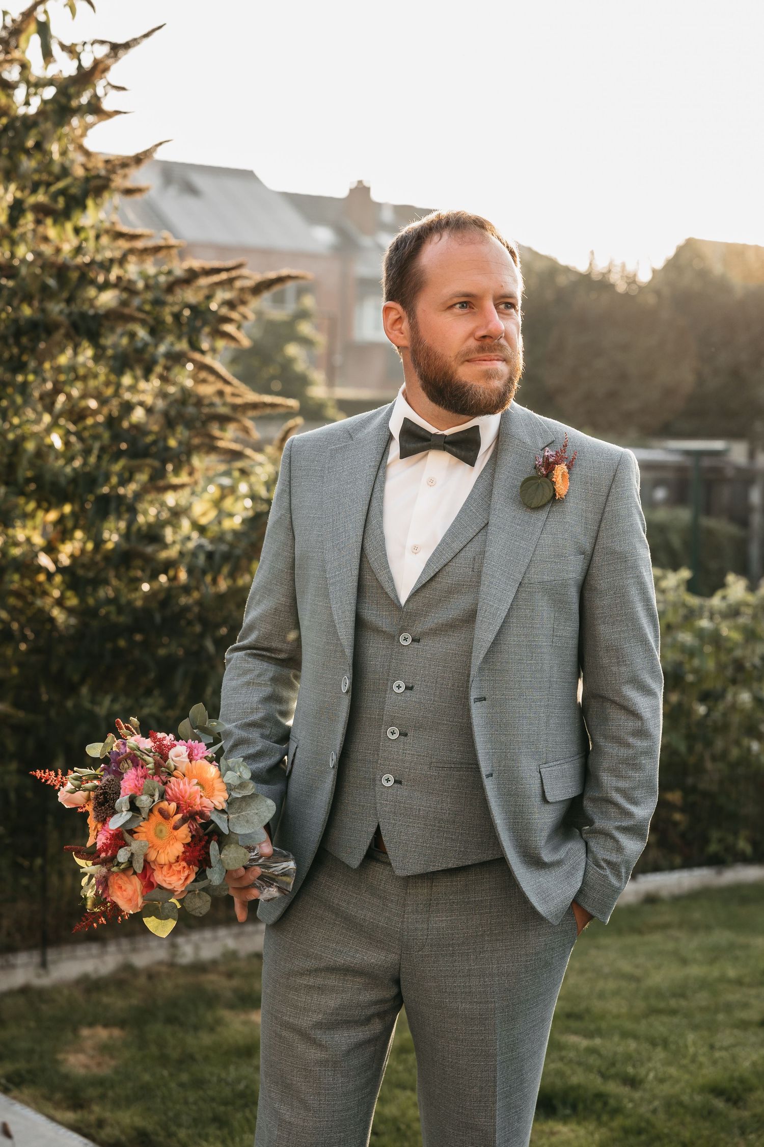 Groom in grey three-piece suit with bow tie holding a colorful bouquet in golden light.