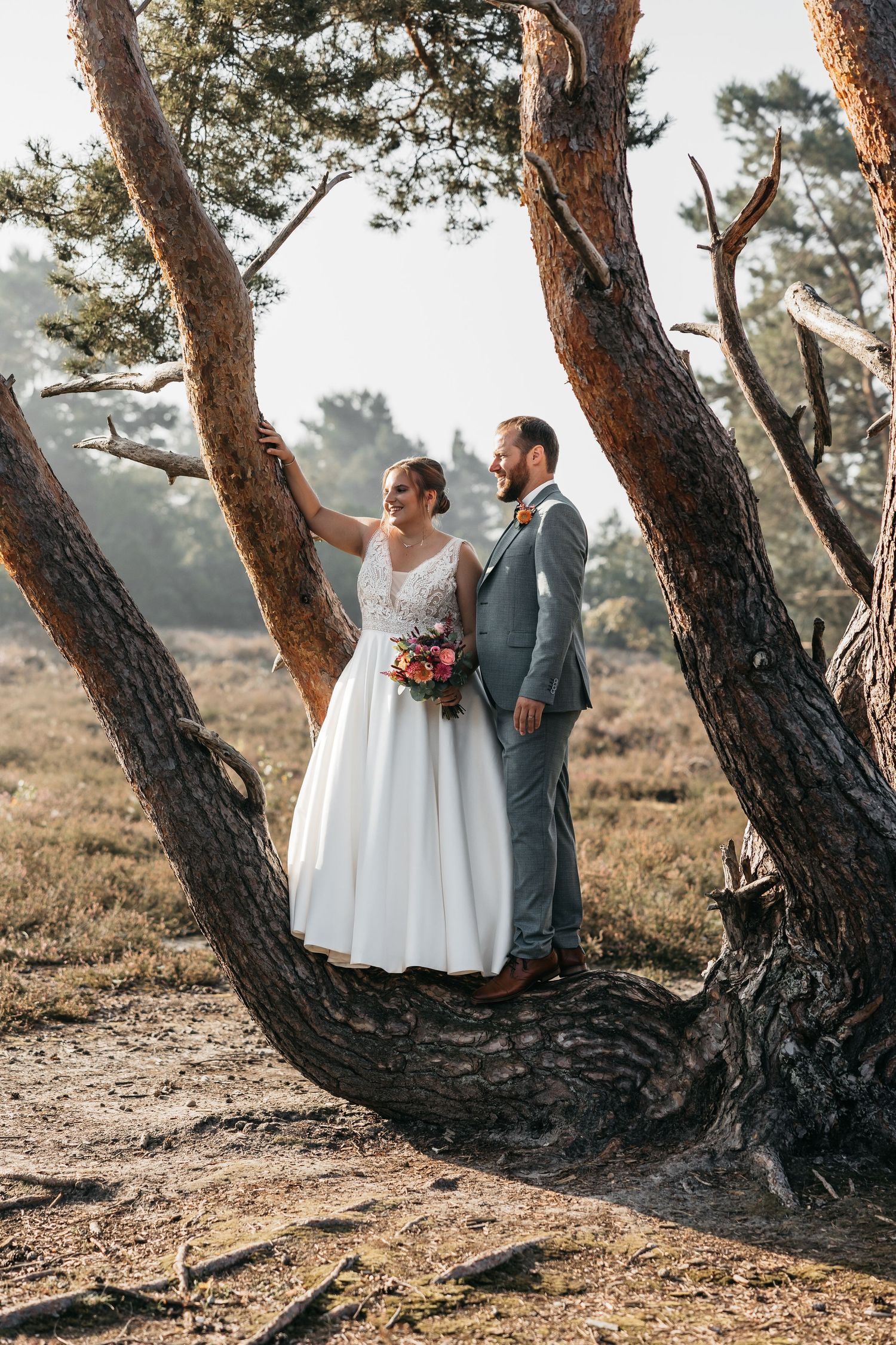 Bride and groom standing together under a curved pine tree in an outdoor heathland setting on their wedding day.