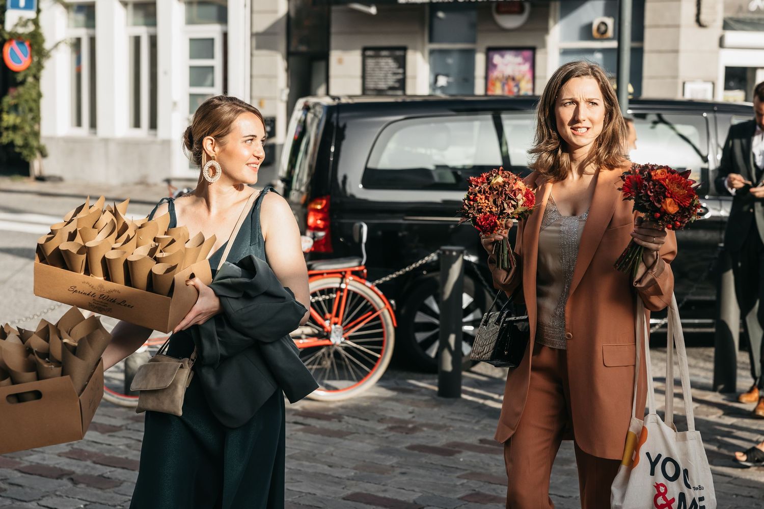 Two women carrying wedding decorations and autumn floral arrangements on a sunny city street.