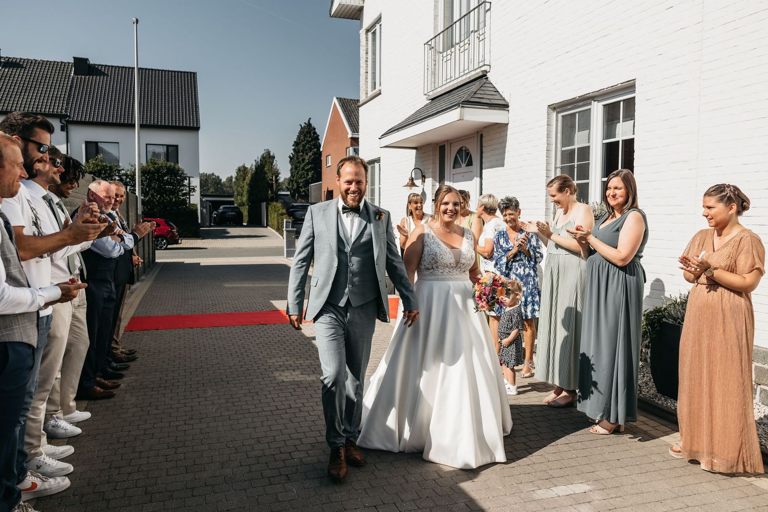 Newlyweds walk triumphantly down a red carpet lined with cheering guests outside a white building.