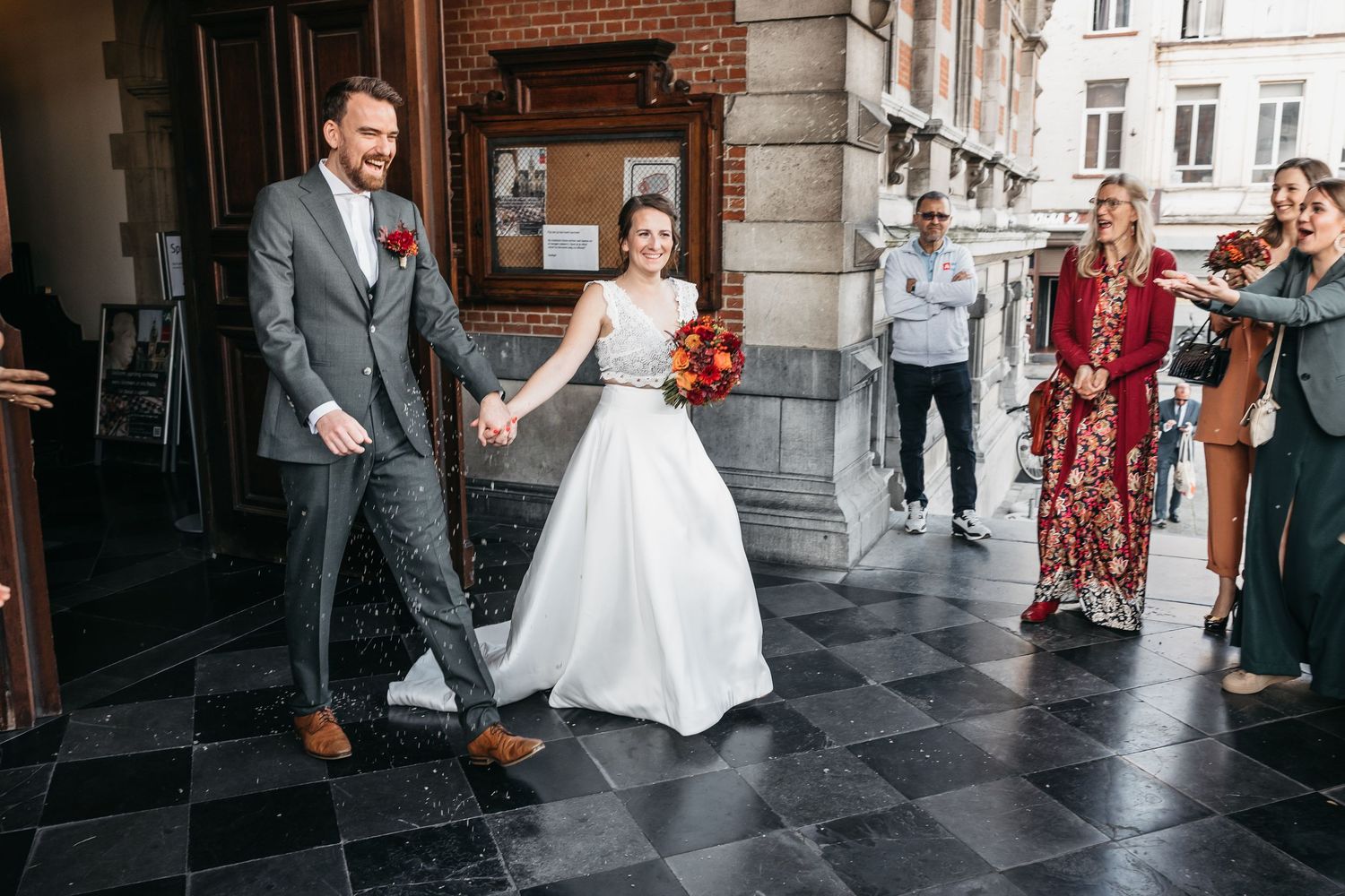 Smiling newlyweds hold hands exiting wedding venue as guests celebrate outside historic building.