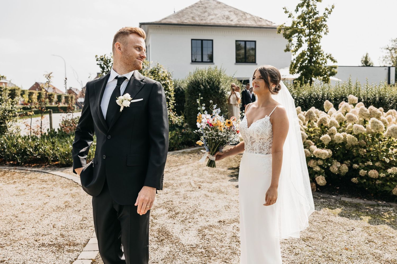 Smiling groom in black suit turns to see bride in lace wedding dress outdoors.