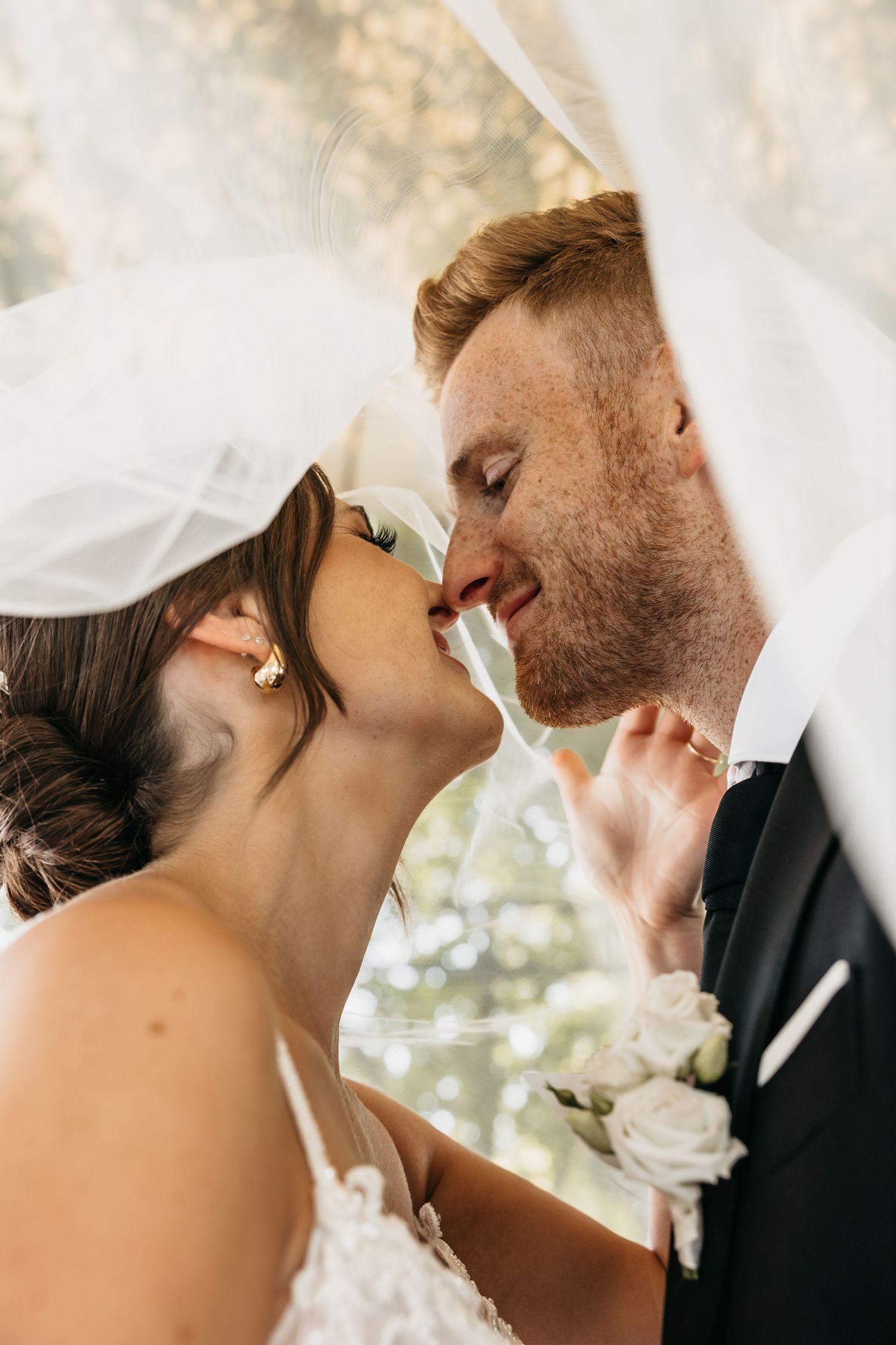 Bride and groom sharing romantic kiss under flowing white veil outdoors.