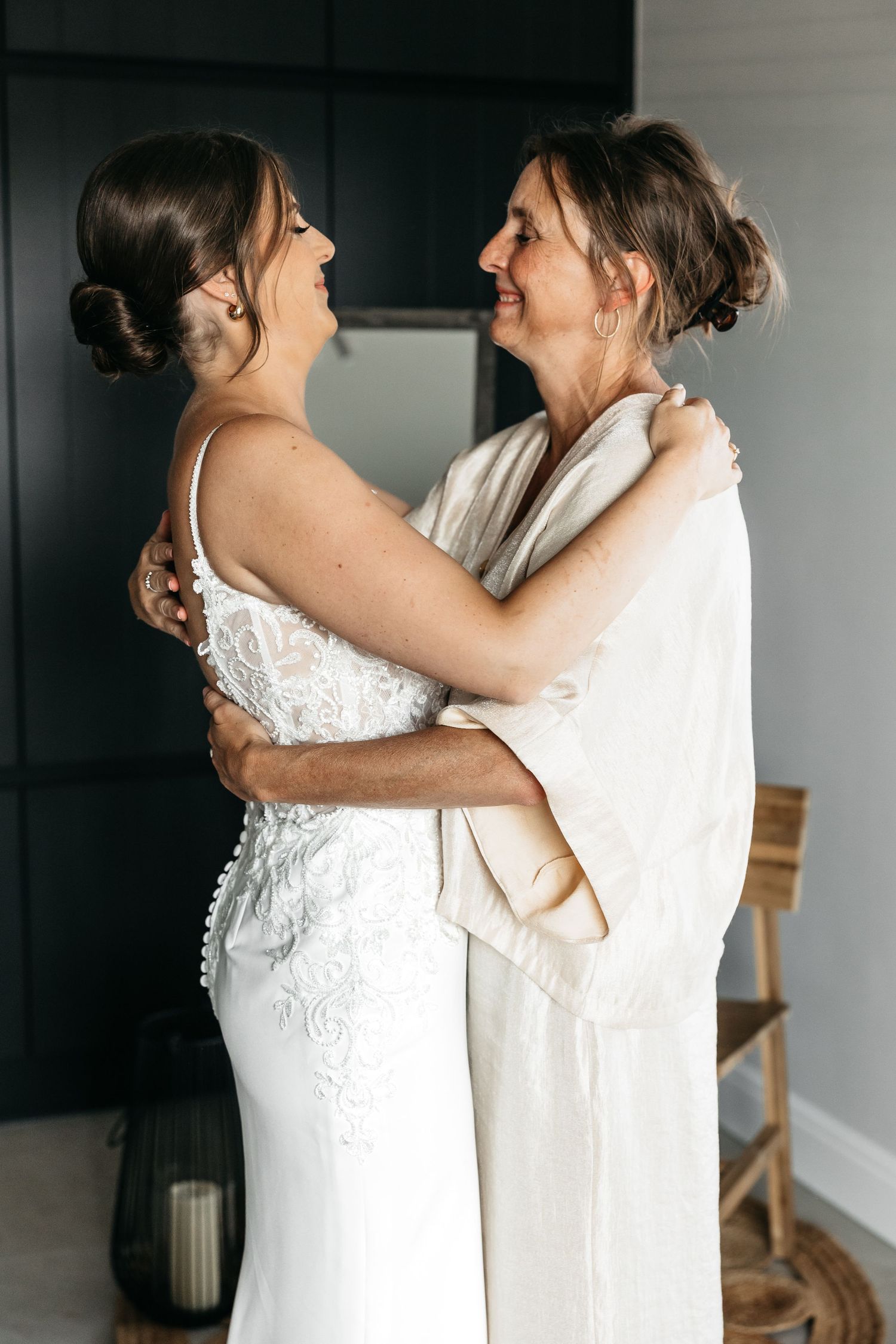 Bride in white lace gown sharing an emotional embrace with woman in white robe.