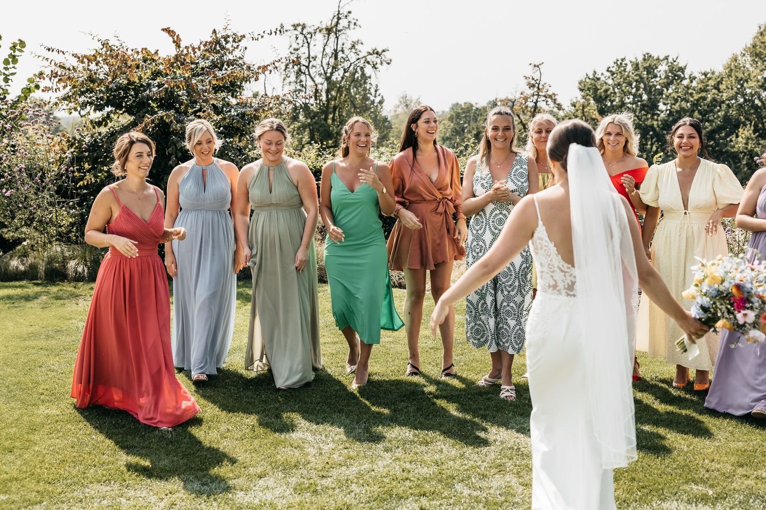 Bride in lace wedding gown swings bouquet toward excited bridesmaids in colorful dresses on sunny garden lawn.