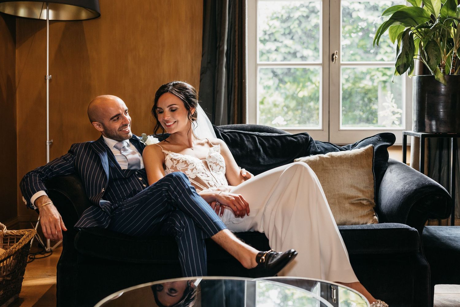 Bride and groom relax together on a dark sofa in a warmly lit elegant interior setting.