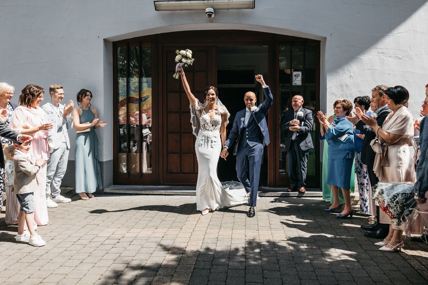 Joyful newlyweds exit wedding venue raising hands in celebration while guests applaud on both sides.