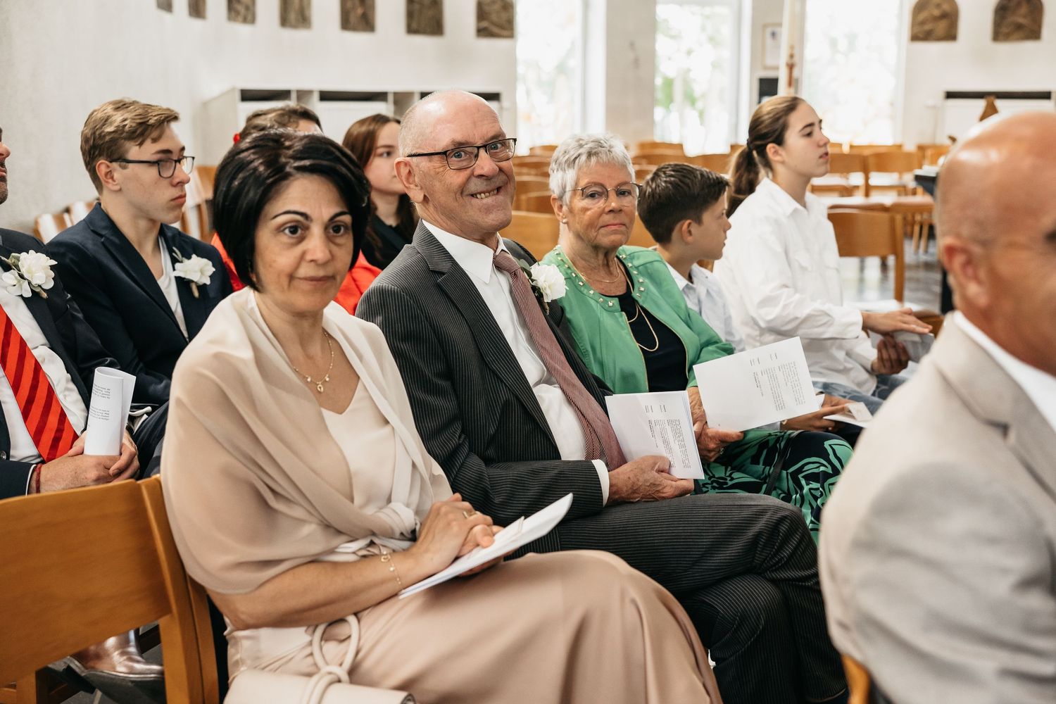 Smiling wedding guests seated in church pews holding ceremony programs during a wedding celebration.
