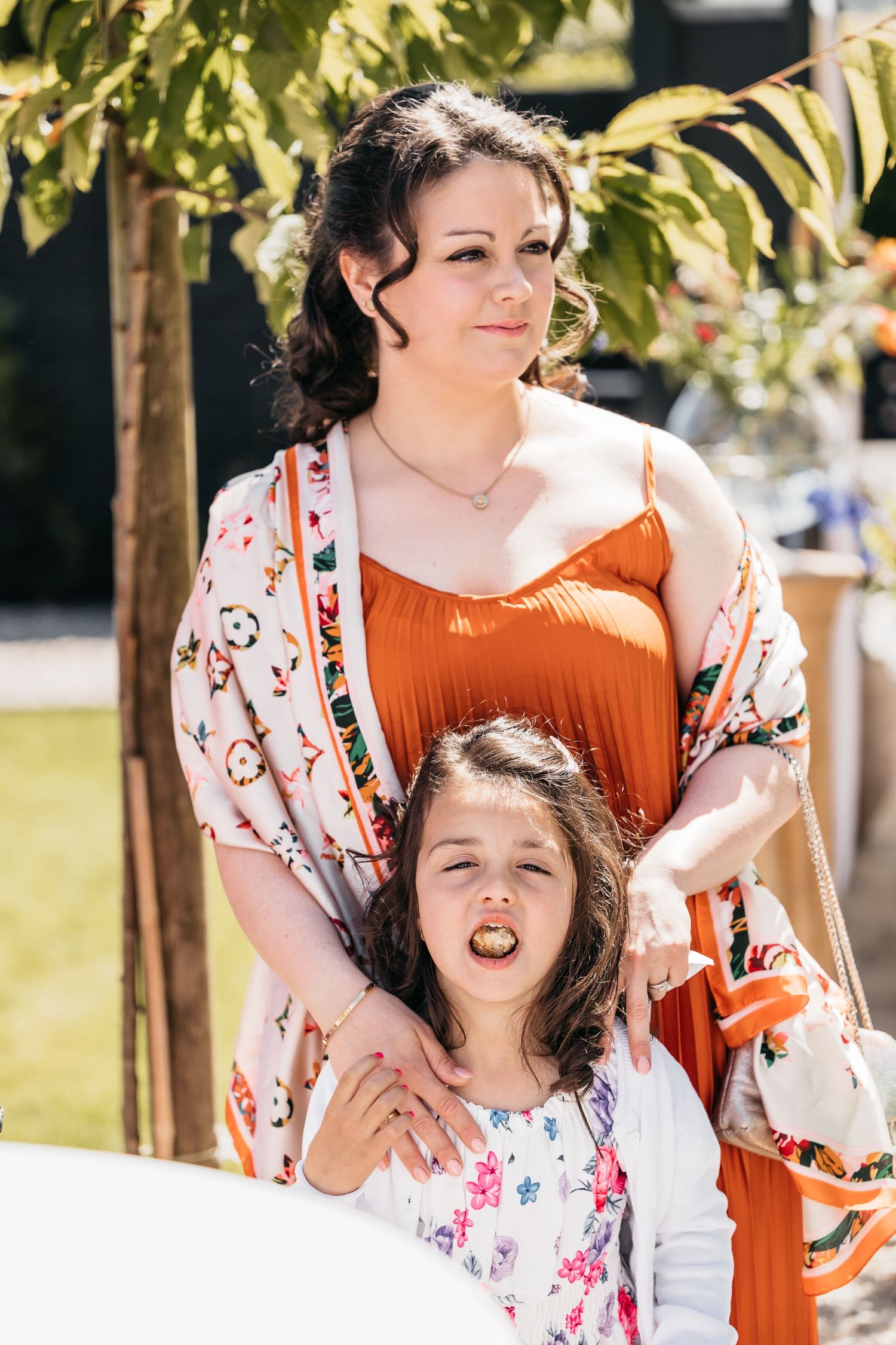 Woman in orange dress and floral kimono standing with young girl at outdoor wedding celebration.