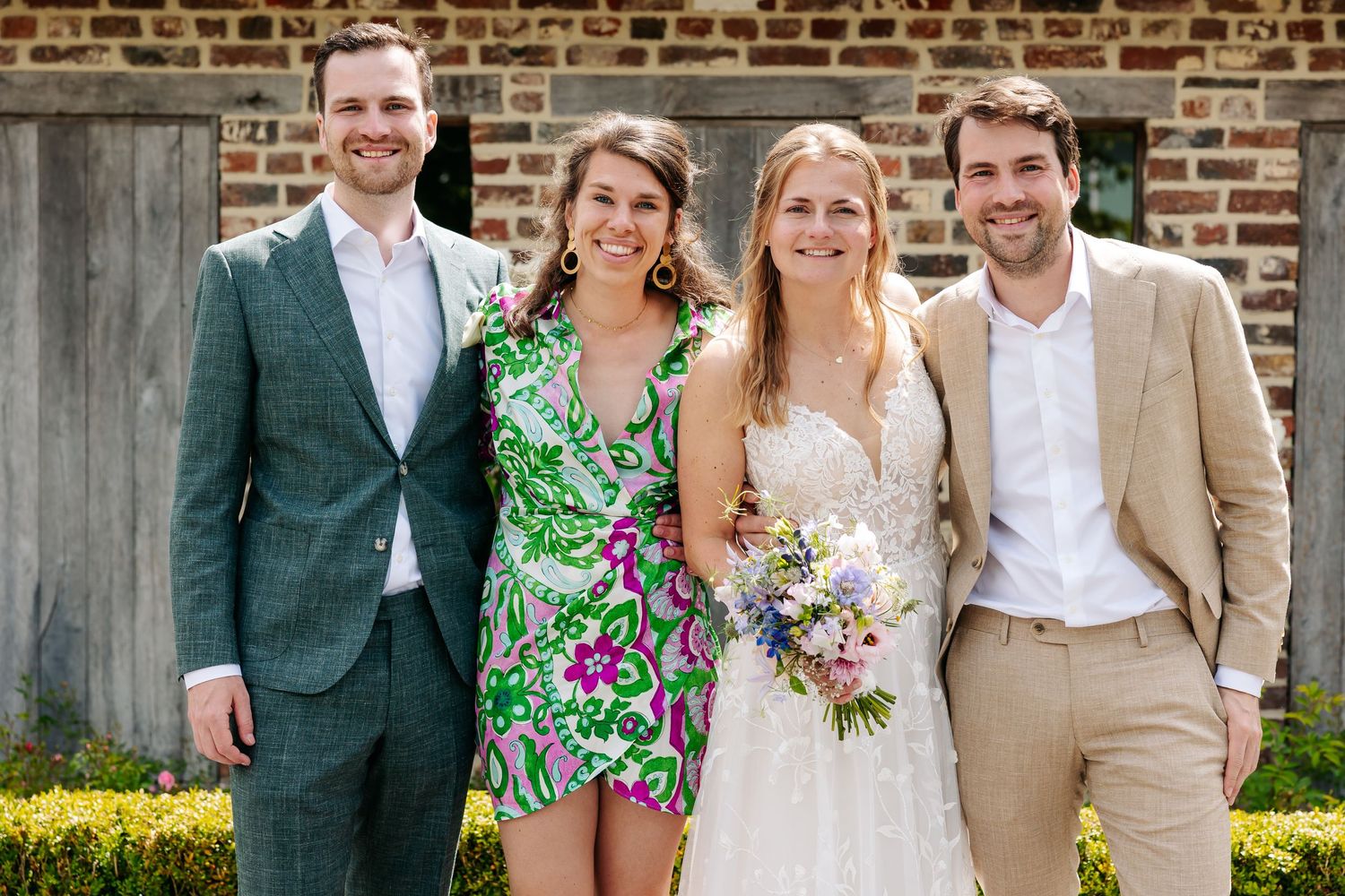 Bride holding wildflower bouquet posing with three guests outside a brick wall at a summer wedding.