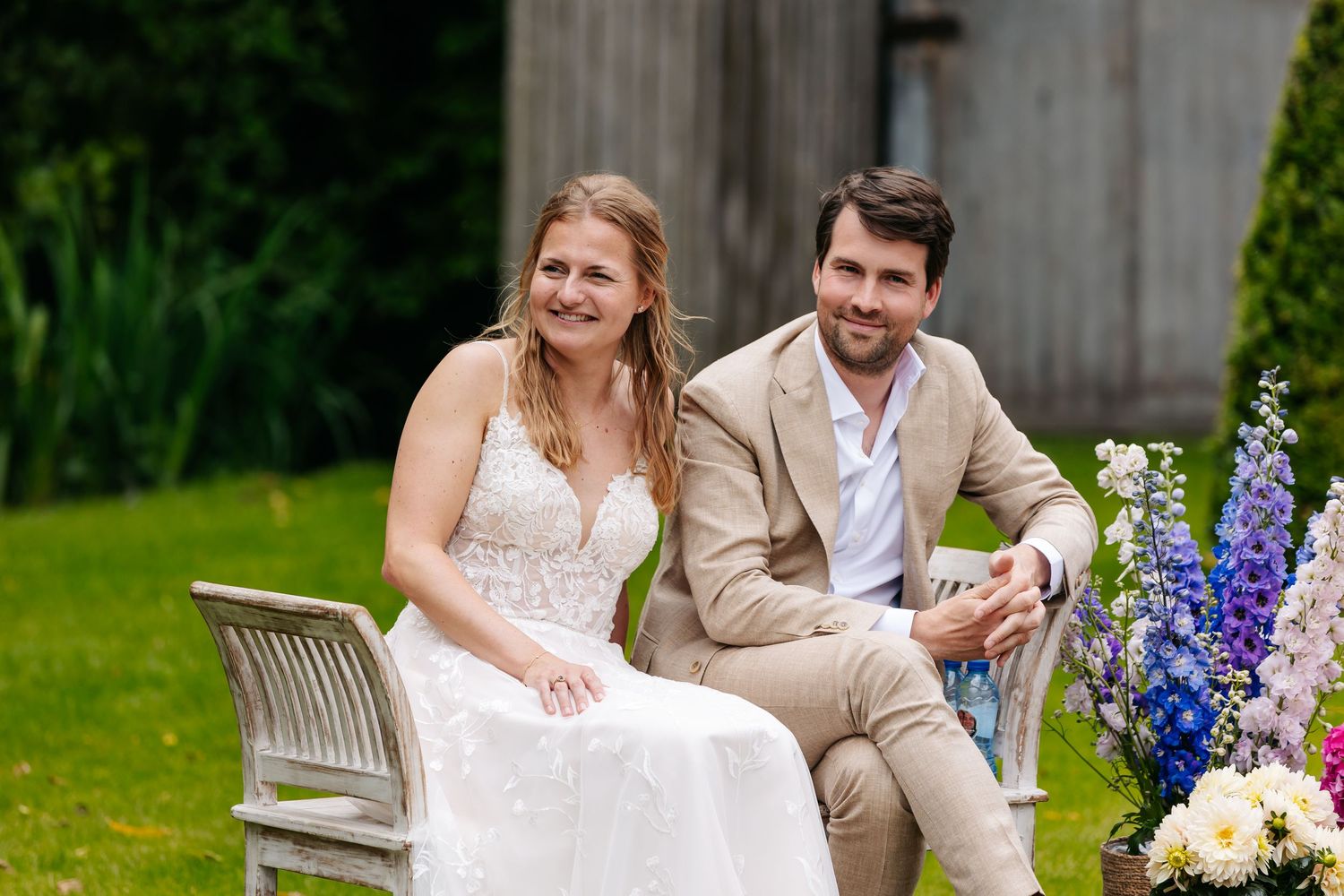 Smiling bride and groom sit together on a garden bench surrounded by colorful flowers.