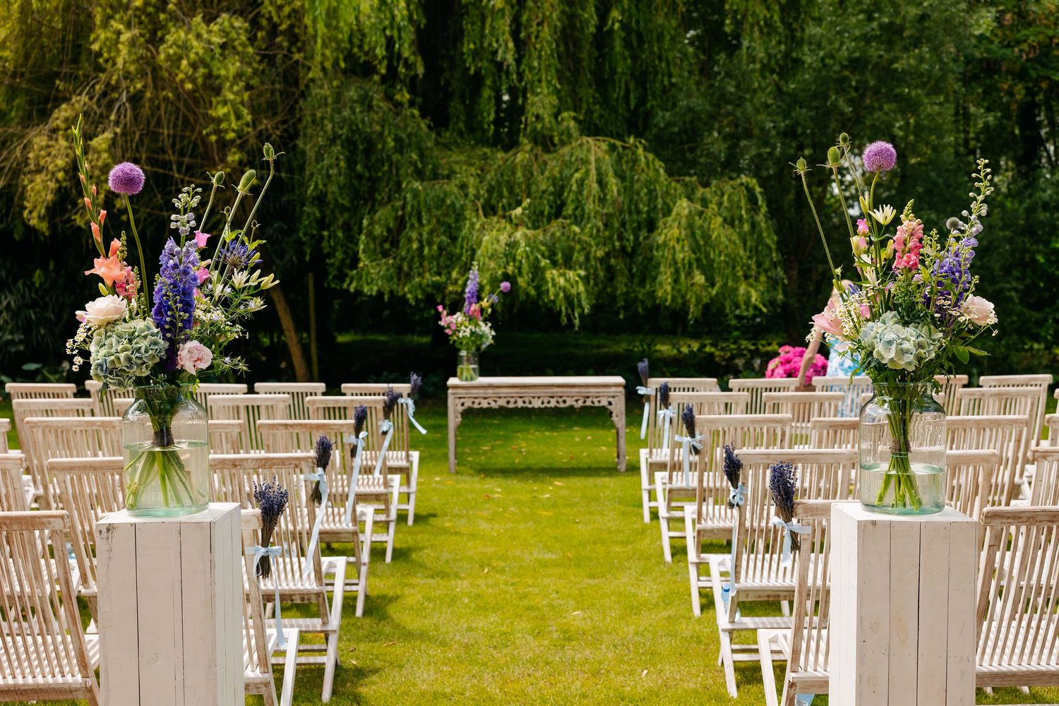 Outdoor wedding ceremony aisle lined with white chairs and tall floral arrangements in a lush green garden setting.