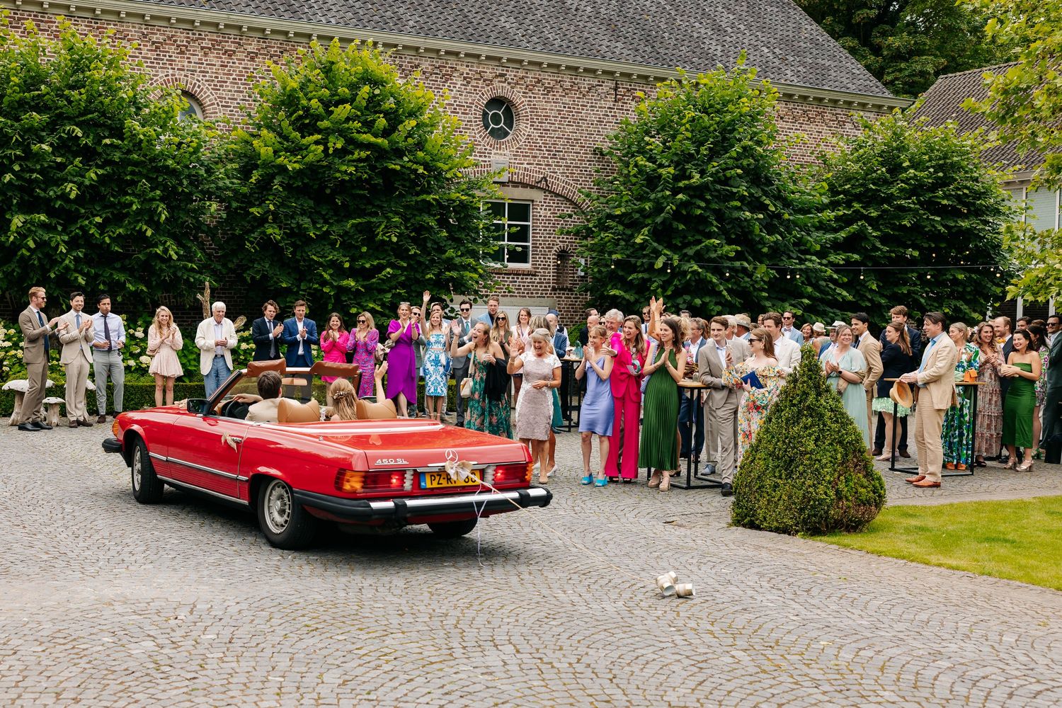 Newlyweds driving away in red convertible Mercedes as wedding guests cheer in courtyard.