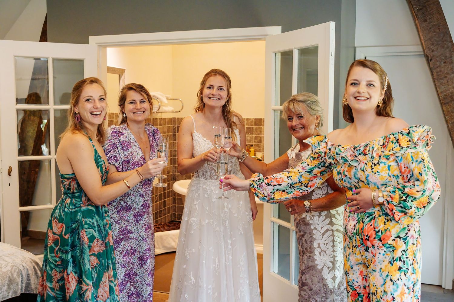 Bride and four bridesmaids in floral dresses toasting with champagne glasses indoors.