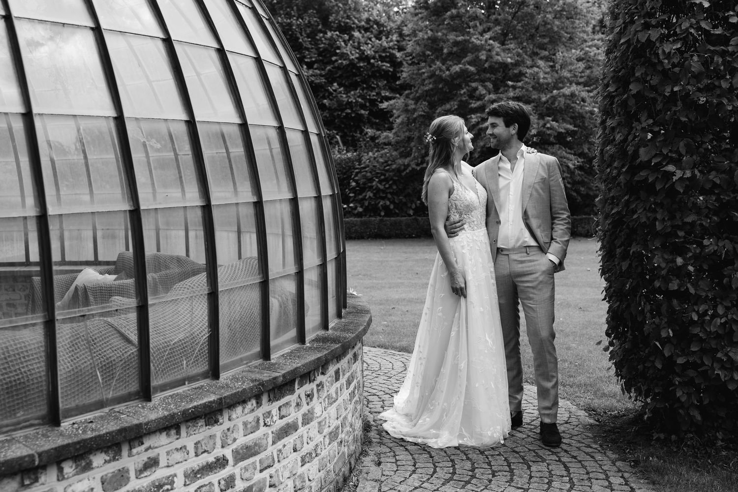 Black and white photo of bride and groom sharing a romantic moment beside a glass greenhouse in a garden.
