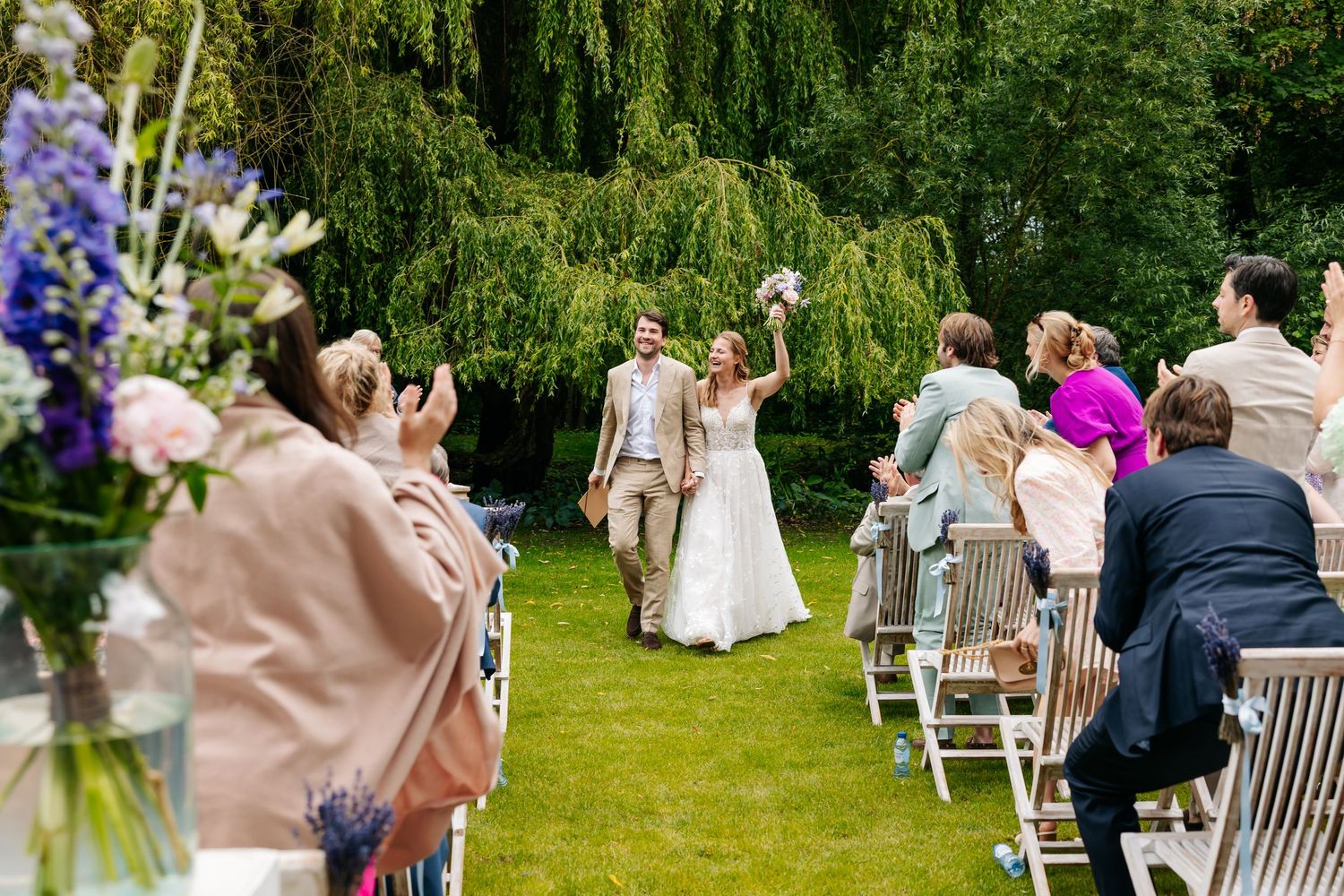 Newlyweds celebrate joyfully walking down aisle as guests applaud at outdoor garden wedding.