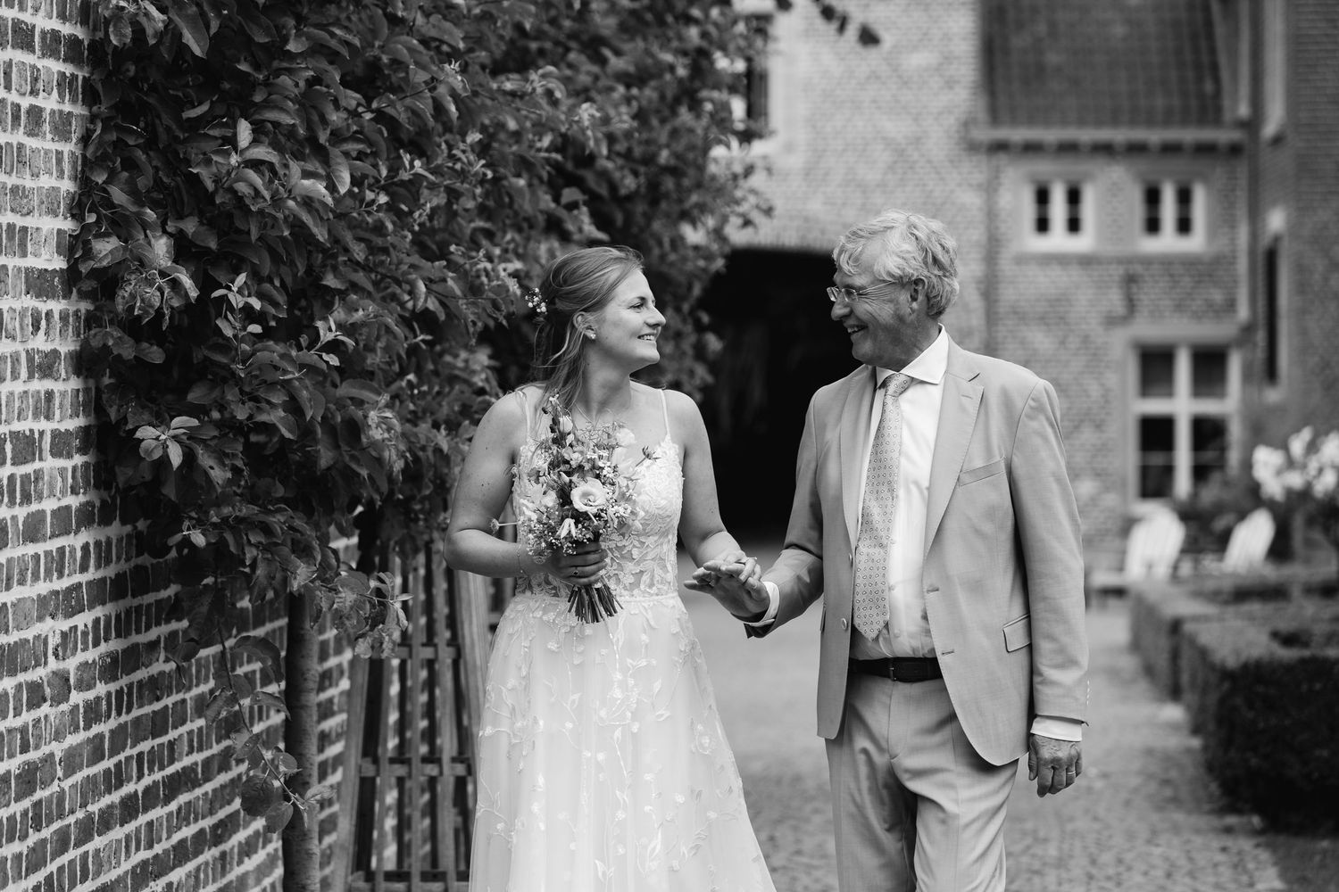 Black and white photo of bride in lace gown holding bouquet walking with older gentleman outside brick building.