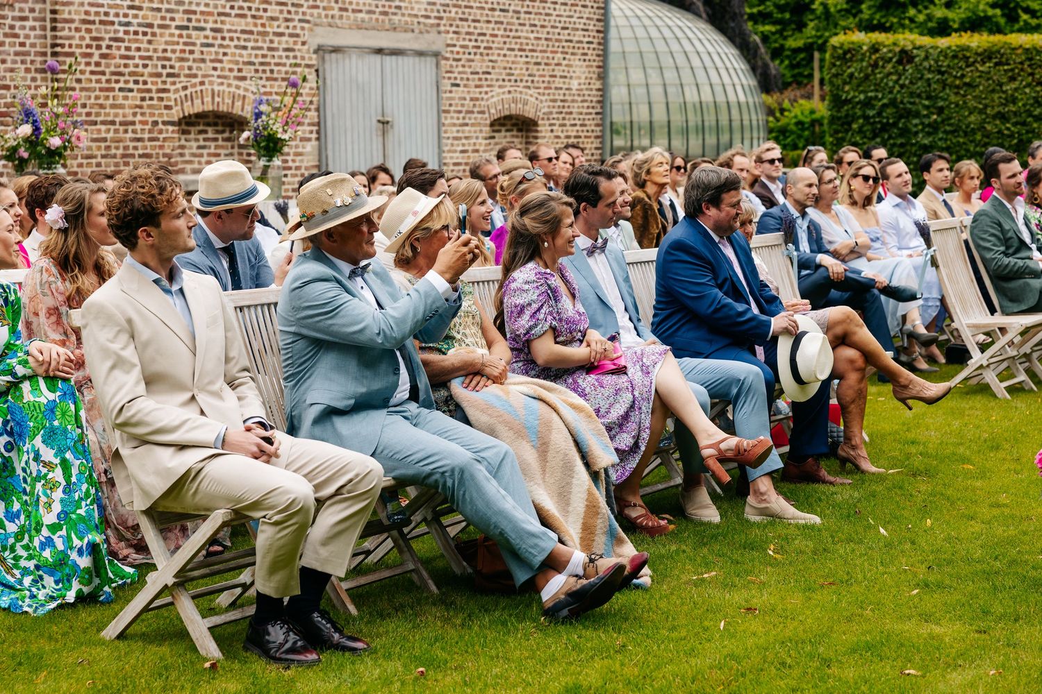 Wedding guests seated outdoors on white chairs enjoying a sunny garden ceremony.