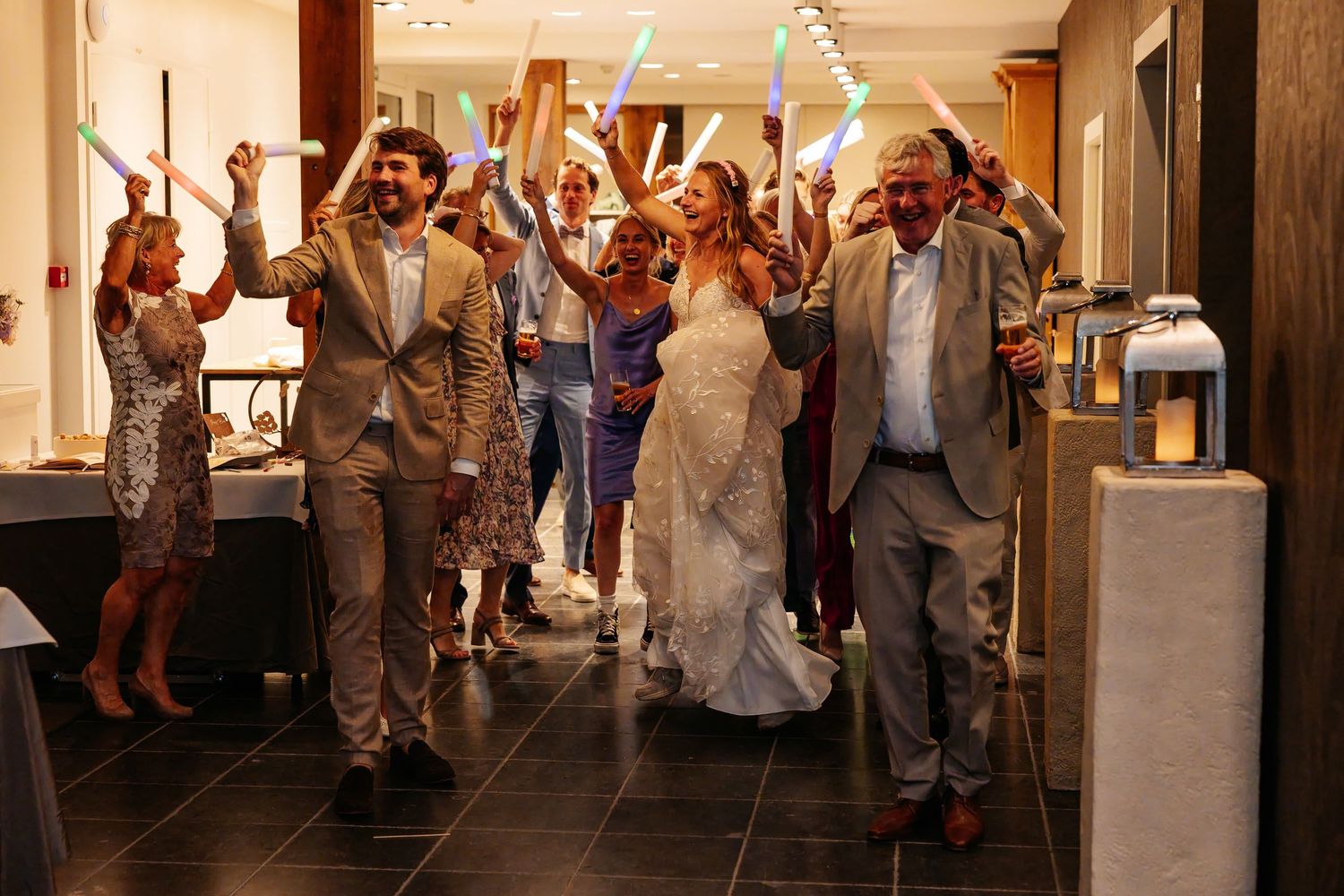 Bride and groom walk through cheering guests waving glow sticks at their festive wedding celebration.