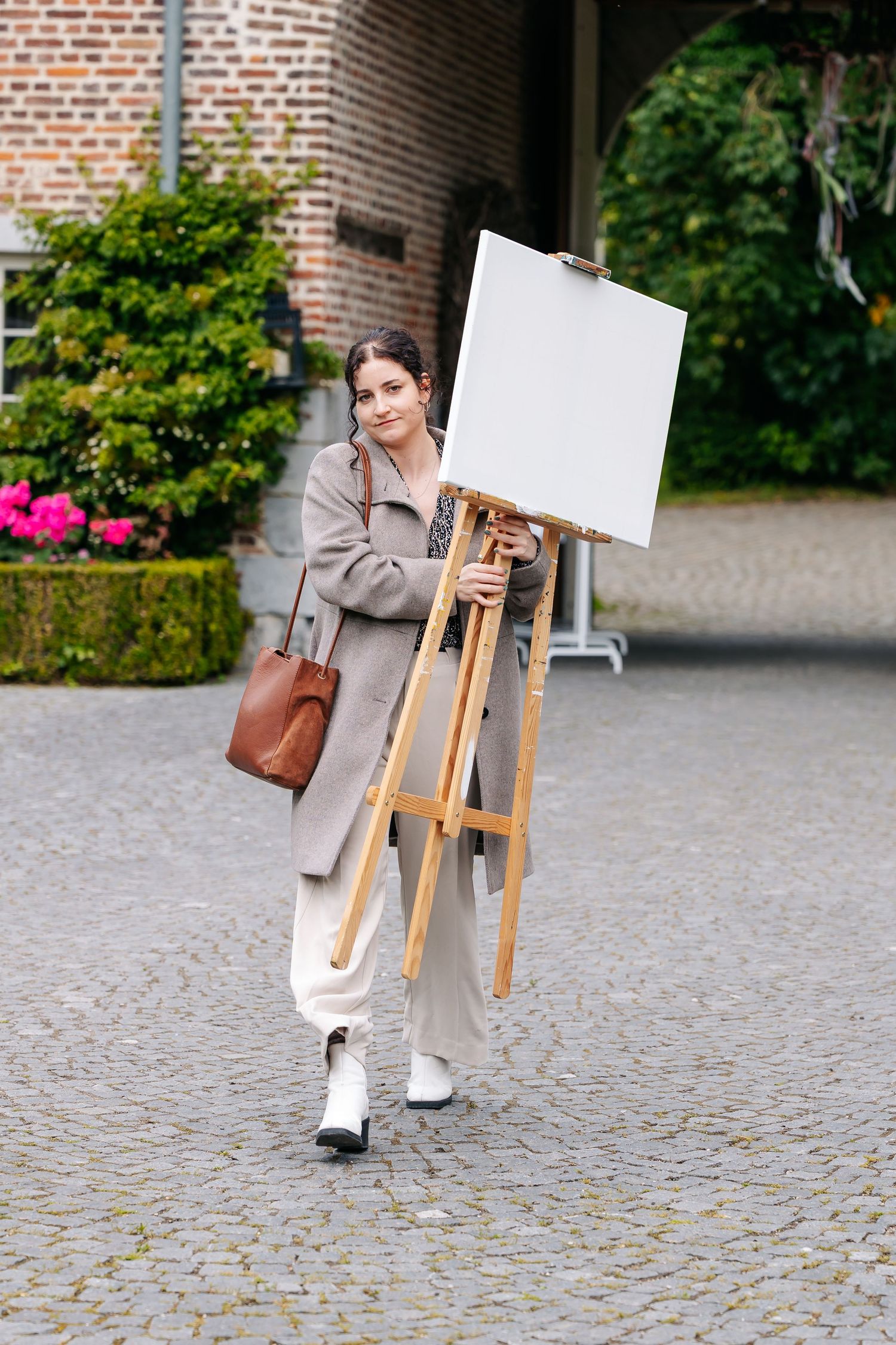 Young woman carrying a wooden easel with blank canvas outdoors on cobblestone courtyard near brick building.