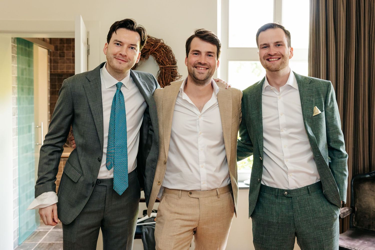 Three smiling groomsmen in suits posing together indoors at a wedding celebration.