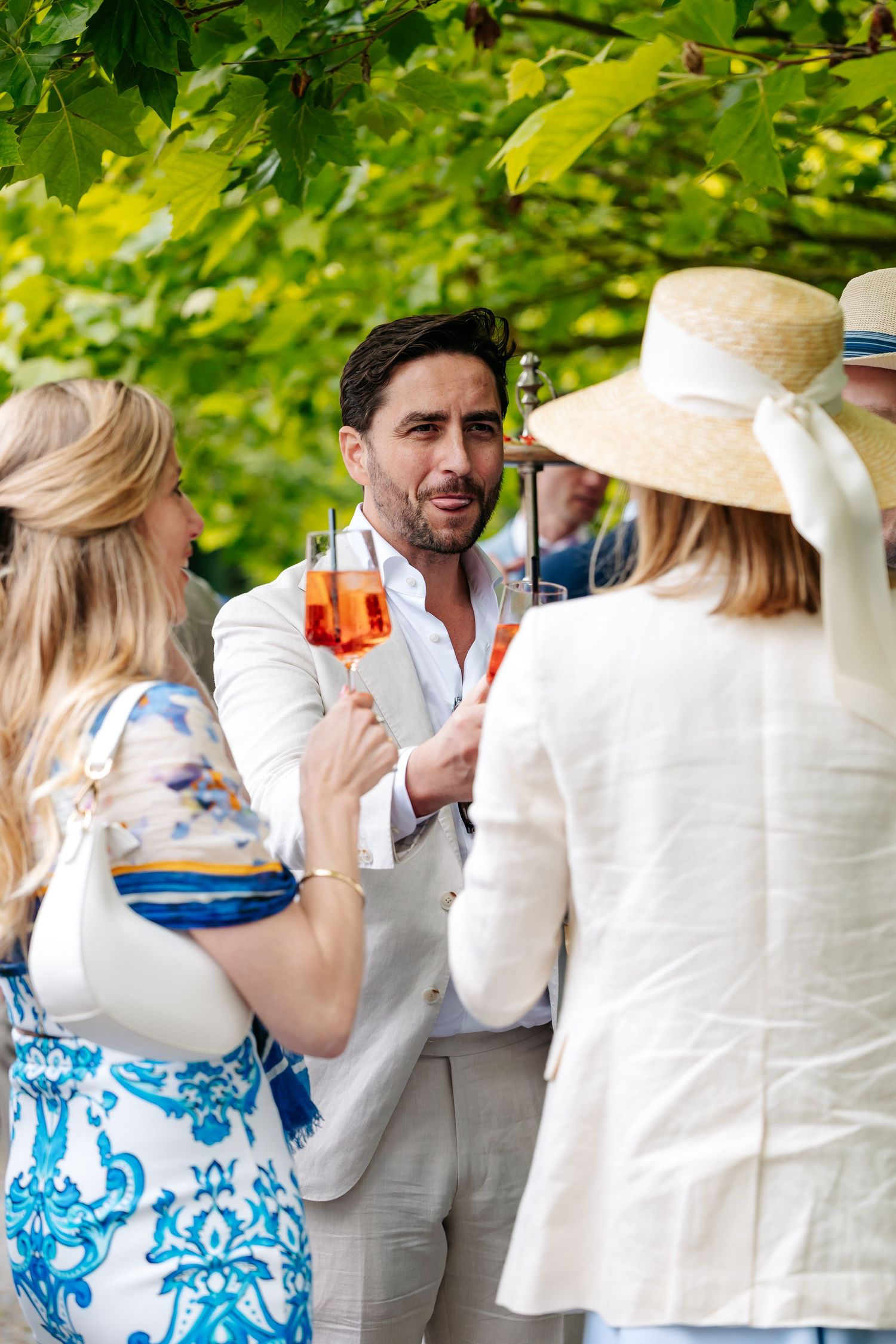 Man in white suit holding aperol spritz conversing with women at outdoor garden party wedding reception.
