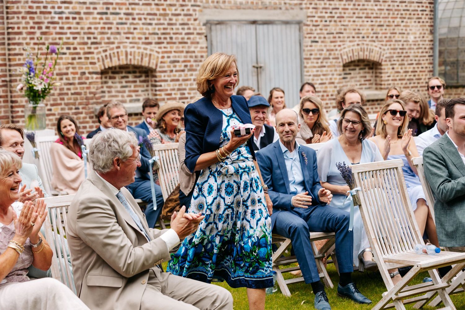 Woman in blue patterned dress speaks at outdoor wedding ceremony while guests seated on wooden chairs applaud.