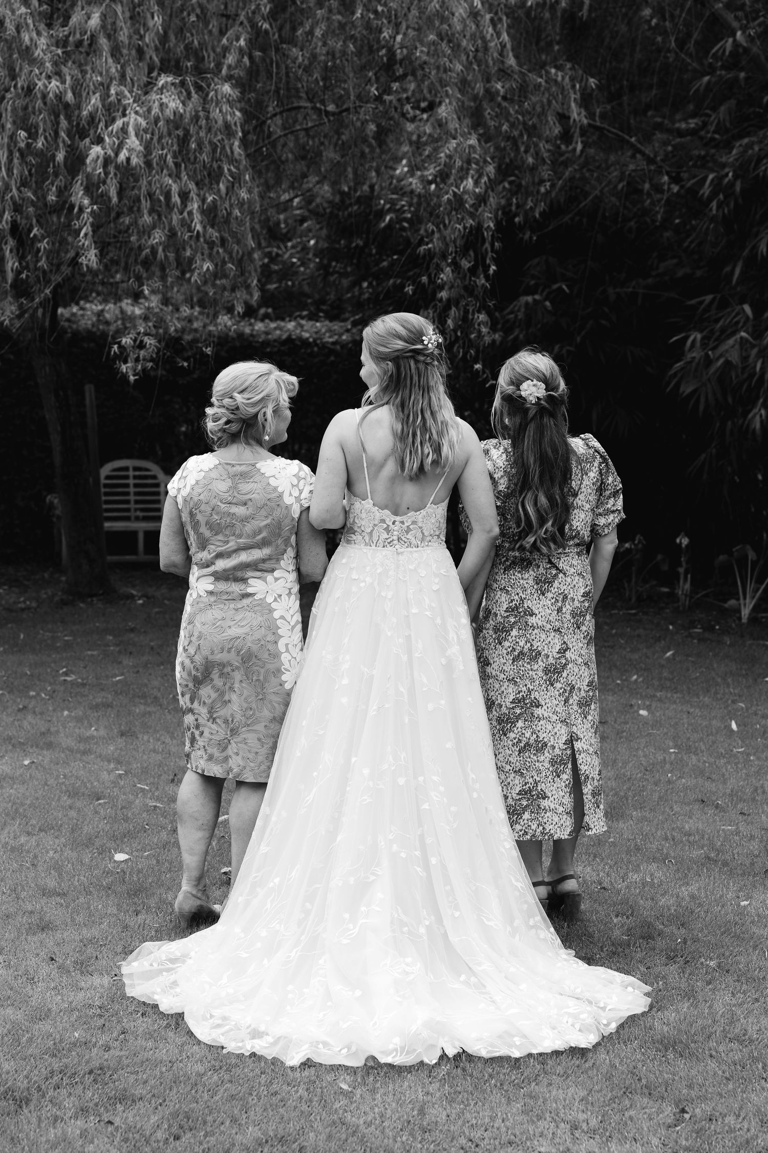 Black and white photo of a bride in a lace gown with two women standing in a garden.