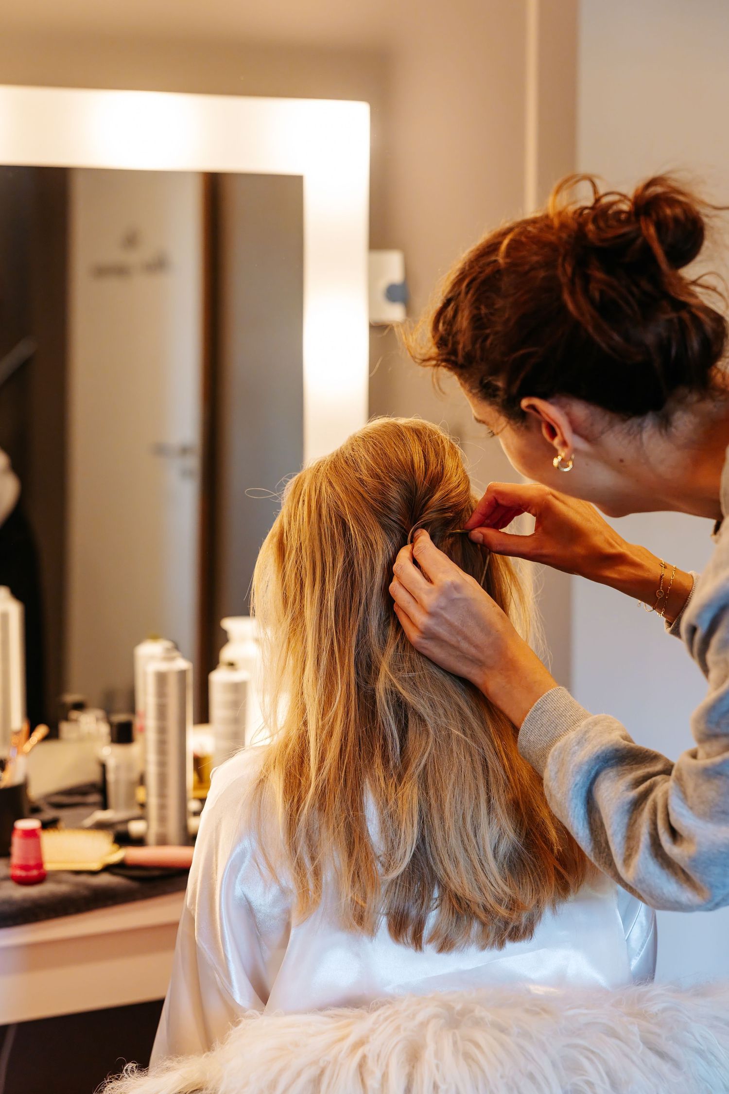 Hairstylist styling blonde hair of bride in front of vanity mirror in bridal preparation room.