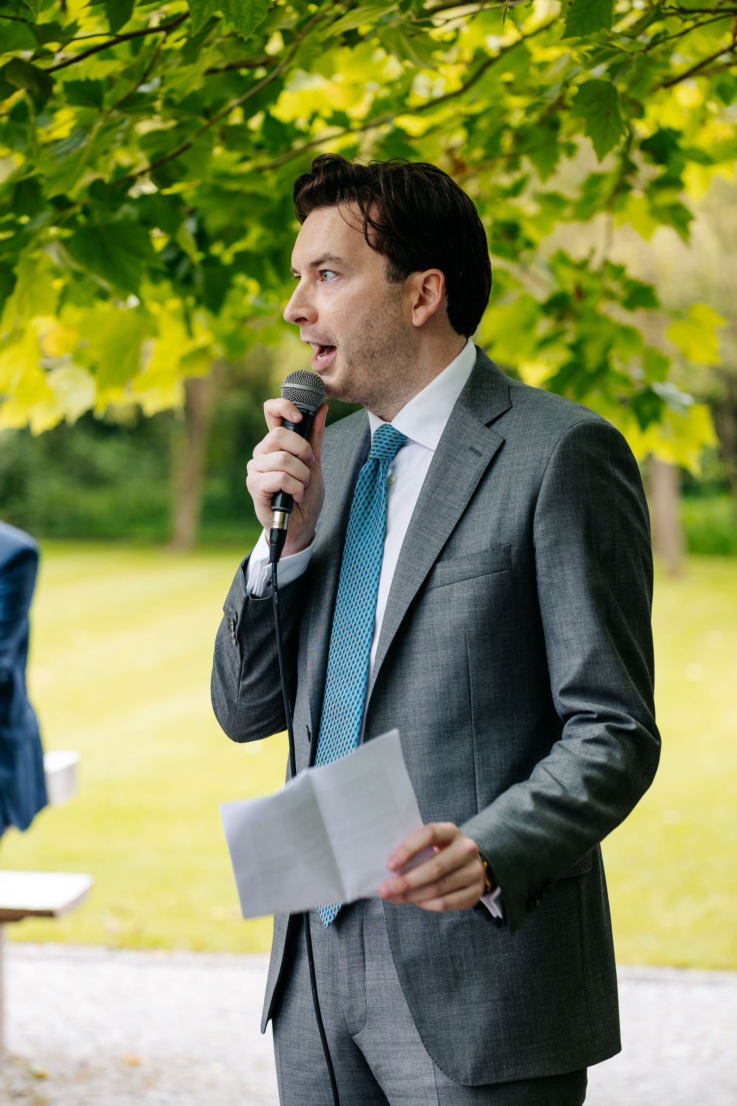 Elegant man in gray suit and blue tie speaking into microphone outdoors under green maple tree canopy.