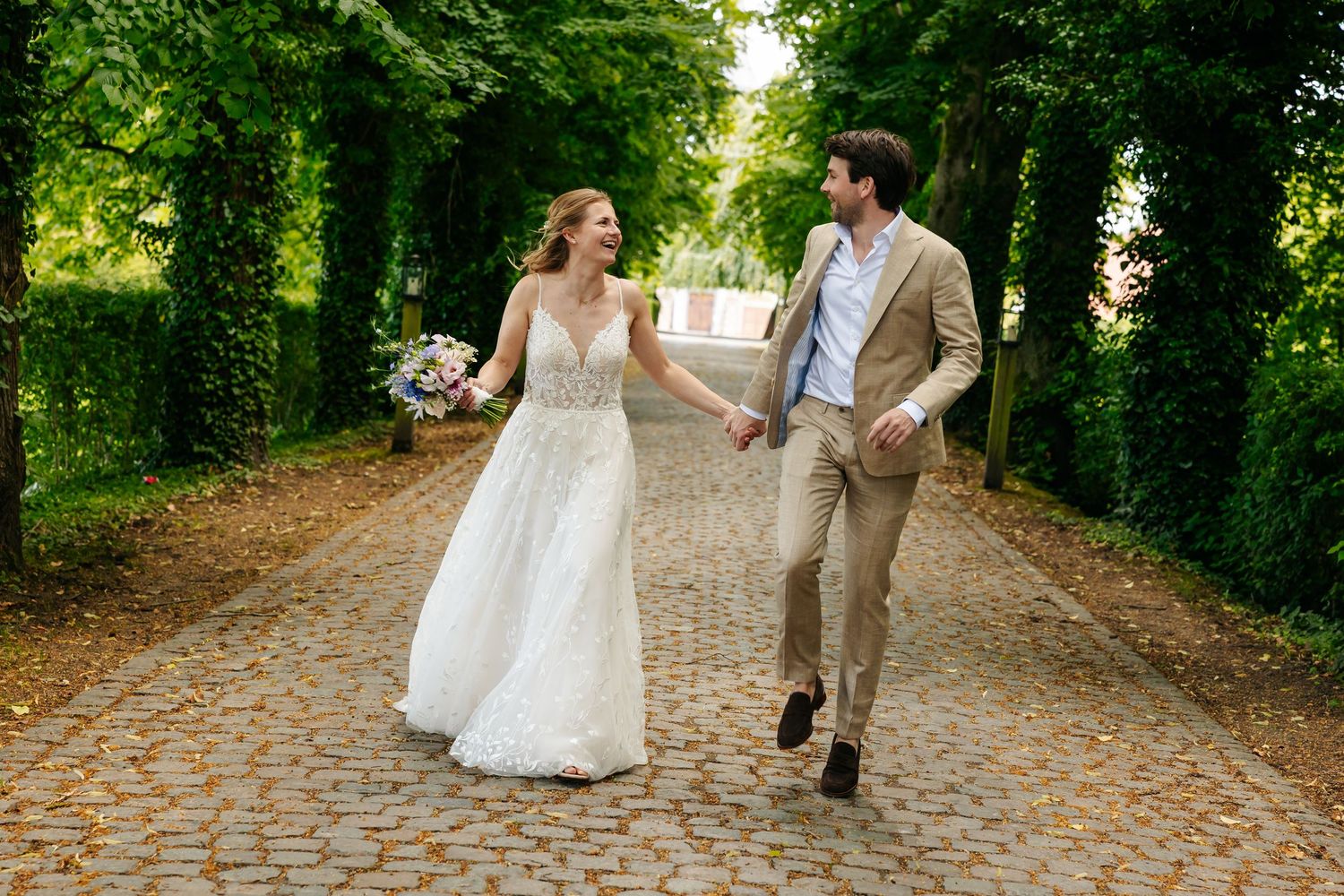 Happy bride and groom walk hand in hand along a lush green tree-lined cobblestone avenue.