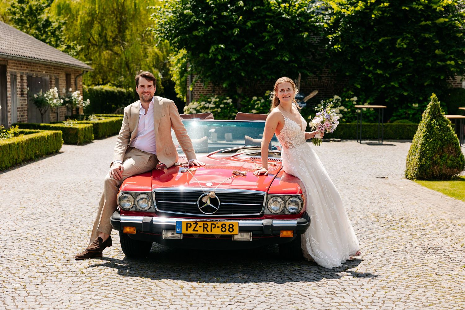 Bride and groom pose leaning against a vintage red Mercedes convertible on cobblestones.