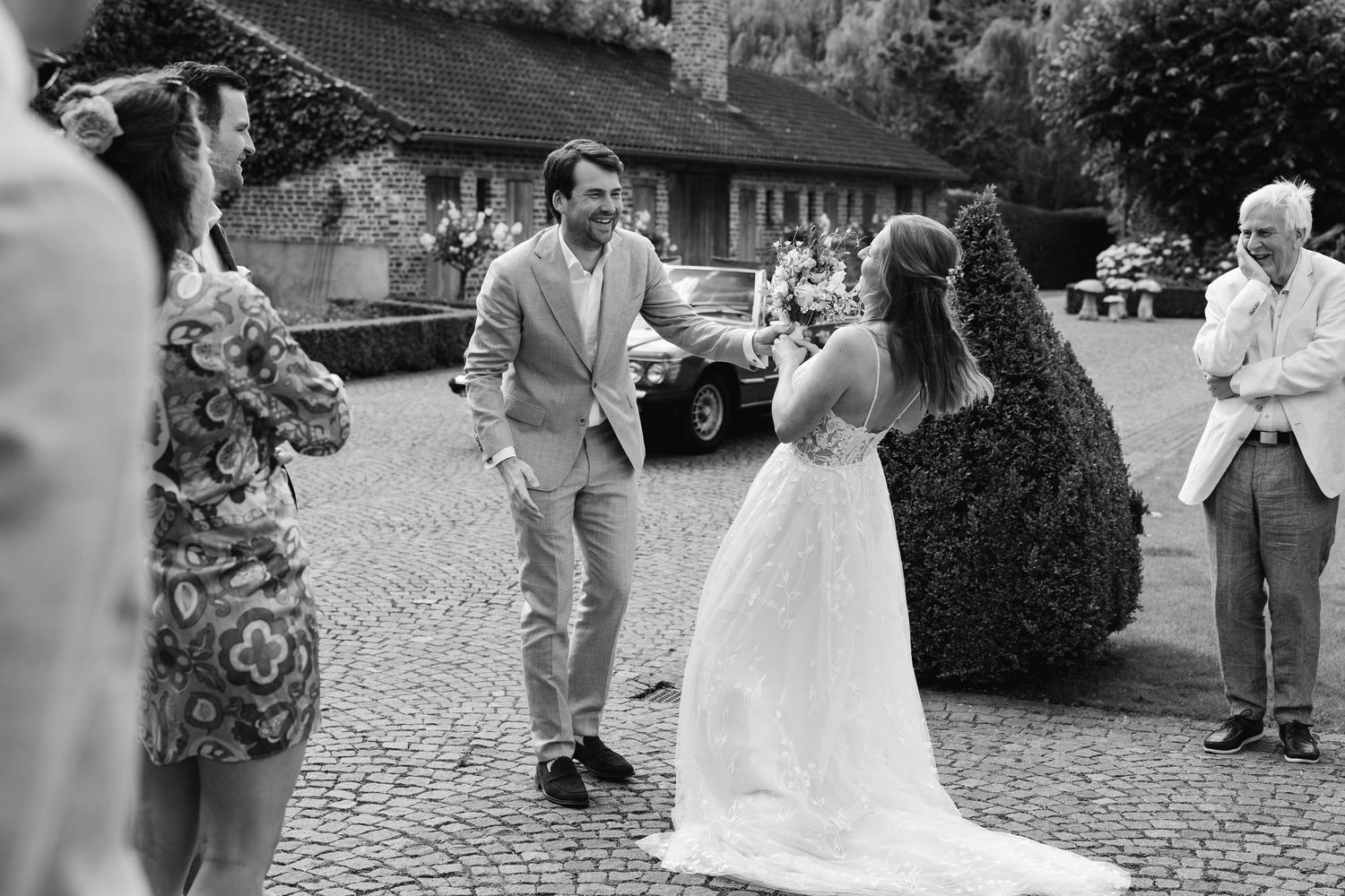Black and white photo of groom approaching bride in wedding dress on cobblestone courtyard.