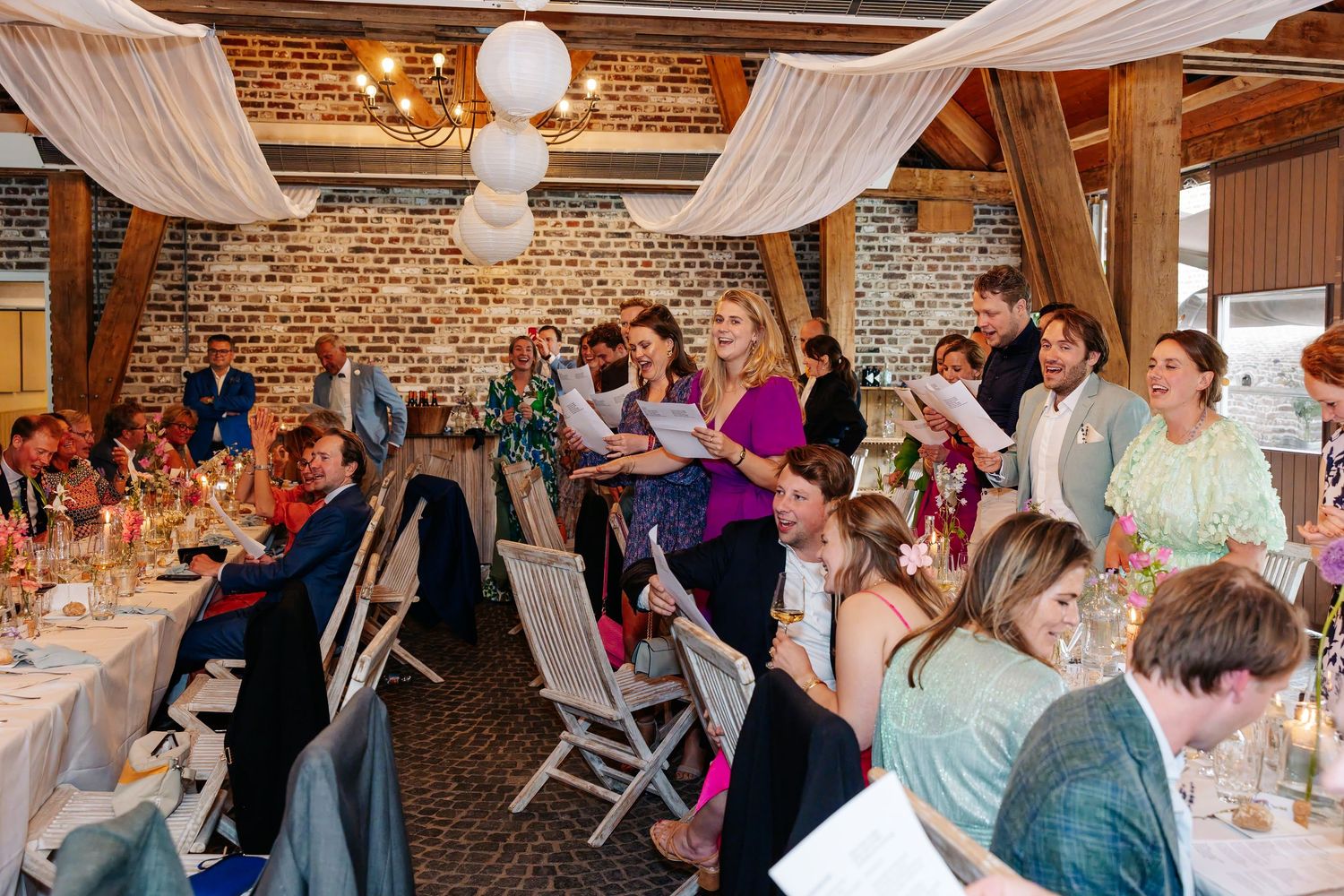 Joyful wedding reception guests laugh and celebrate together at long decorated dinner tables in barn.