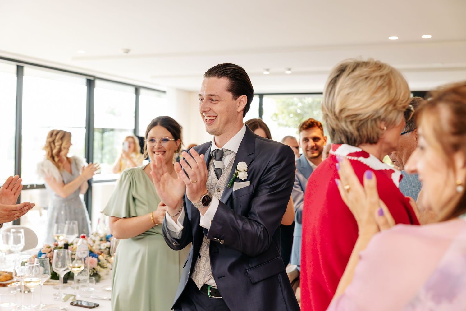 Joyful groom in navy suit laughs and claps with guests during lively wedding reception celebration.